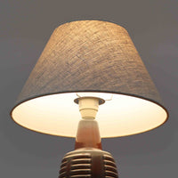 Rigmor Nielsenがデザインし、Soholm Keramikが手掛けたテーブルランプです。高さ37cmの取り入れやすいサイズに、艶やかな陶磁器ベースとナチュラルなリネンシェードを組み合わせた、落ち着きある北欧ビンテージらしい一台。ベースには規則的な立体リブ装飾が施され、陰影豊かな表情と彫刻的な存在感を生み出しています。点灯時にはシェード越しの柔らかな光が釉薬の奥行きや貫入の表情を引き立て、チェストやサイドテーブル、ベッドサイドに穏やかな雰囲気を演出。Rigmor Nielsenらしい造形美とSoholmの高い陶芸技術が感じられる、希少性の高いビンテージ照明です。