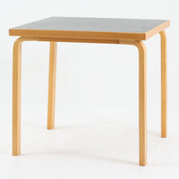 Alvar Aaltoがデザインし、Artekより製作された「81C」ダイニングテーブルです。バーチ材を用いたフレームに、天板には落ち着いた質感のリノリウムを組み合わせたシンプルで機能的なデザインが特徴。脚部にはアアルトを象徴するL字型の曲木構造が採用されており、軽やかで柔らかな印象を与えます。幅75cmの正方形サイズはダイニングはもちろんワークテーブルとしても使いやすく、日常の空間に北欧デザインの美しさと実用性をもたらすビンテージ家具です。本個体は、脚部の5枚レイヤー構造、1973年以降に採用されたプラスネジ、および天板裏の3段構成スタンプの仕様から、1973年~1990年代始めに掛けて製造されたと思われます。