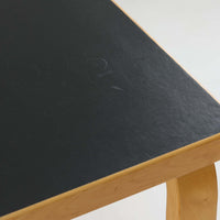 Alvar Aaltoがデザインし、Artekより製作された「81C」ダイニングテーブルです。バーチ材を用いたフレームに、天板には落ち着いた質感のリノリウムを組み合わせたシンプルで機能的なデザインが特徴。脚部にはアアルトを象徴するL字型の曲木構造が採用されており、軽やかで柔らかな印象を与えます。幅75cmの正方形サイズはダイニングはもちろんワークテーブルとしても使いやすく、日常の空間に北欧デザインの美しさと実用性をもたらすビンテージ家具です。本個体は、脚部の5枚レイヤー構造、1973年以降に採用されたプラスネジ、および天板裏の3段構成スタンプの仕様から、1973年~1990年代始めに掛けて製造されたと思われます。