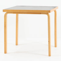 Alvar Aaltoがデザインし、Artekより製作された「81C」ダイニングテーブルです。バーチ材を用いたフレームに、天板には落ち着いた質感のリノリウムを組み合わせたシンプルで機能的なデザインが特徴。脚部にはアアルトを象徴するL字型の曲木構造が採用されており、軽やかで柔らかな印象を与えます。幅75cmの正方形サイズはダイニングはもちろんワークテーブルとしても使いやすく、日常の空間に北欧デザインの美しさと実用性をもたらすビンテージ家具です。本個体は、脚部の5枚レイヤー構造、1973年以降に採用されたプラスネジ、および天板裏の3段構成スタンプの仕様から、1973年~1990年代始めに掛けて製造されたと思われます。