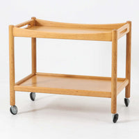 Hans J. Wegnerがデザインし、PP Moblerが製造したAT45のキャスターワゴンです。明るく温かなオーク材を用い、上下2段の棚板と四隅のキャスターを組み合わせた、移動性に優れた実用的な構成が特徴。上段には持ち手を兼ねる開口部が設けられ、緩やかに湾曲する側板や丸みのある支柱など、ウェグナーらしい柔らかな造形と丁寧な木工技術が感じられます。経年によって深まった木肌を楽しみながら、キッチンやダイニング、リビングで配膳や収納に活躍する一台です。
