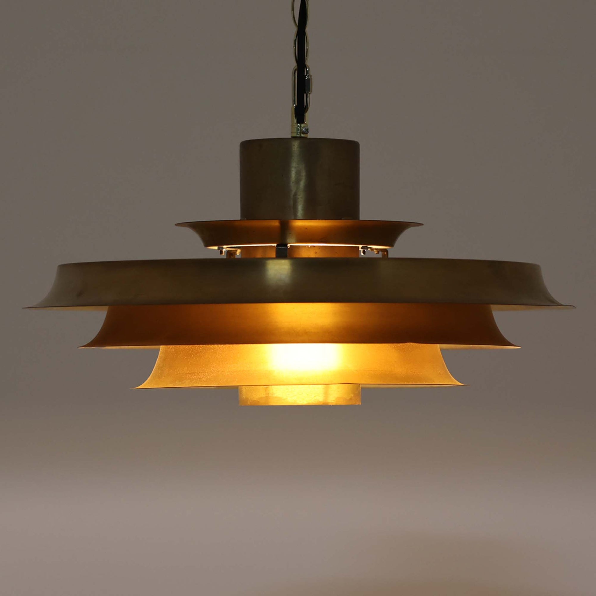 波打つように重なったシェードが印象的なデンマーク製のペンダントライト。無垢の真鍮によるしっかりとした重量感と、内側から放たれる柔らかな光が調和し、空間に温かみと品格を添えます。経年によって色味変化した真鍮は、この照明の歴史と個性を物語り、北欧ビンテージらしい味わい深い魅力を一層引き立てます。ダイニングやリビングのアクセントとして最適な一灯です。