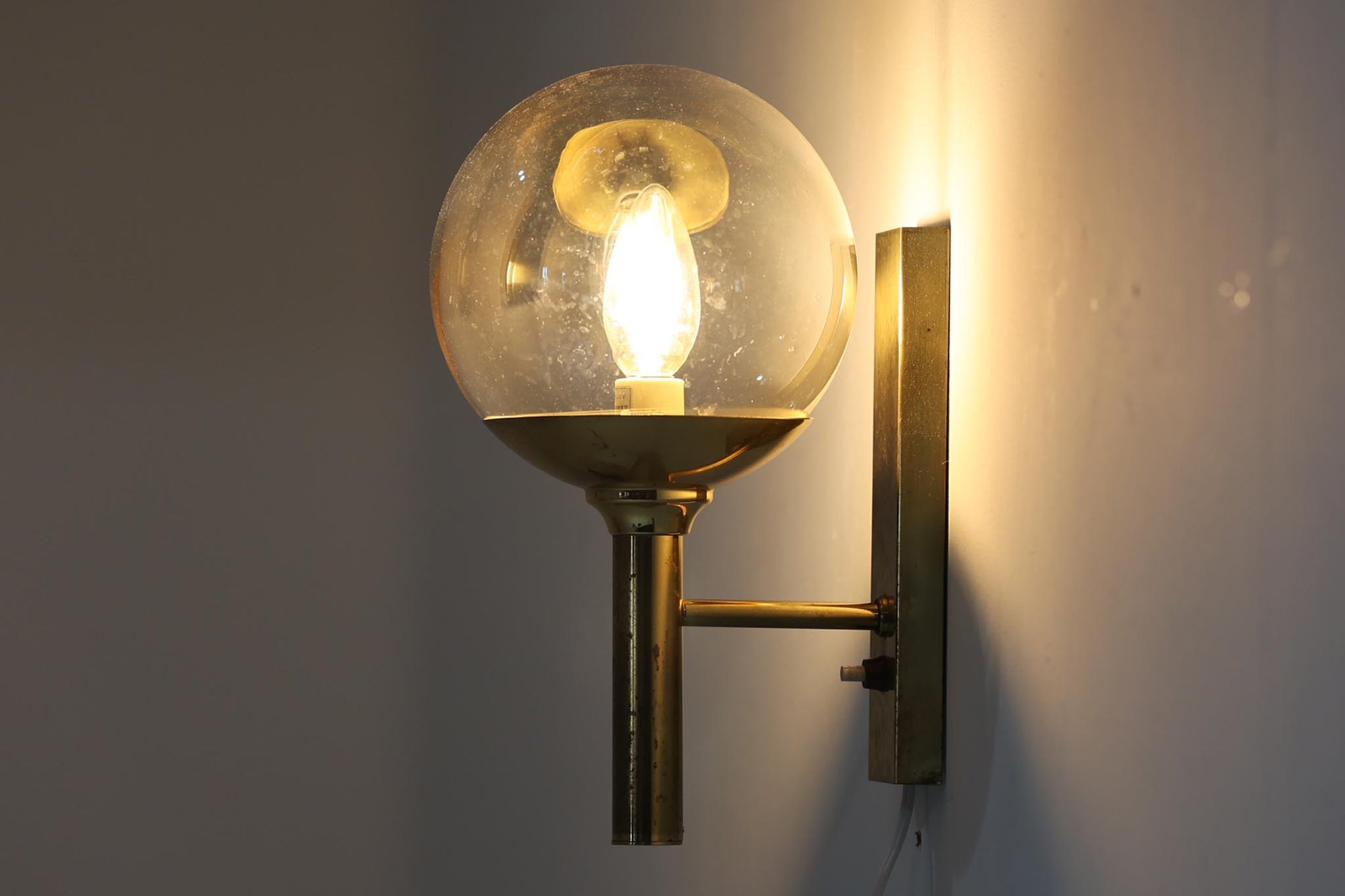こちらは、Sven mejlstromによってデザインされた1960年代のウォールランプです。Sven mejlstromは、ノルウェーの照明デザイナーとして知られ、シンプルで機能的な美しさを追求したデザインを手掛けました。このランプは、真鍮とクリアガラスを組み合わせた美しいデザインが特徴で、ビンテージ品ならではの風合いが感じられます。大きなボール状のガラスシェードは光を柔らかく拡散し、空間に温かみと上品さを加えます。経年変化による真鍮部分の風合いも、時間を経た美しさを感じさせる魅力的な要素です。シンプルでありながらも印象的なこのウォールランプは、どんなインテリアにも調和し、エレガントなアクセントを与えてくれる一品です。