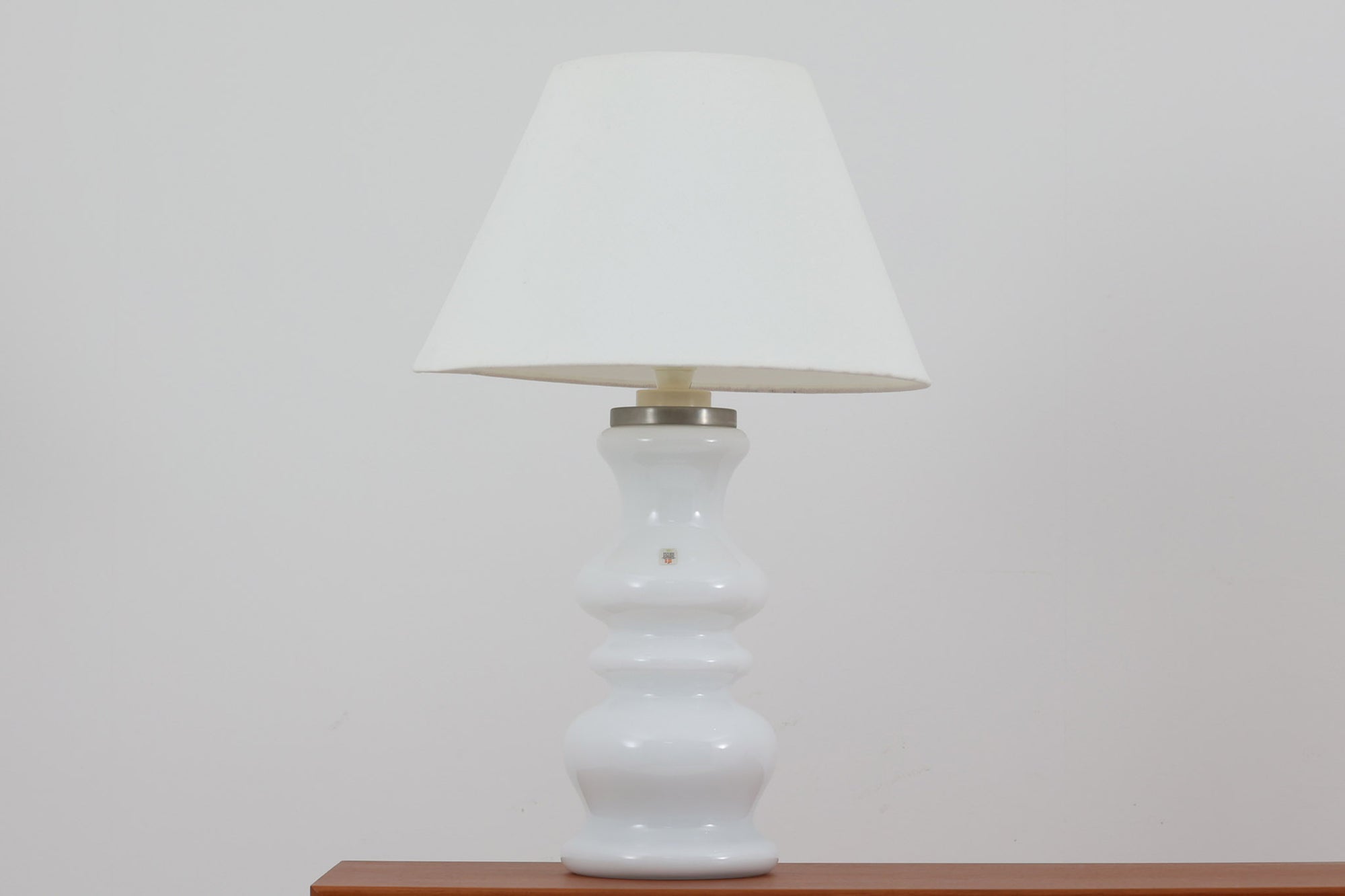 著名なデザイナー、Sidse Werner（シセ・ヴェルナー）による「BIBLIOTEK」のテーブルランプです。ペンダントランプはよく見かけますが、テーブルランプは非常に珍しいモデルです。ホルムガードならではの厚みのある高品質なガラスを使用しており、点灯するとシェードを通して柔らかな温かみのある光が広がり、空間を優雅に照らします。高さ64cmの大型サイズで存在感があり、リビングや書斎、ベッドサイドなど、あらゆるインテリアに美しく調和します。