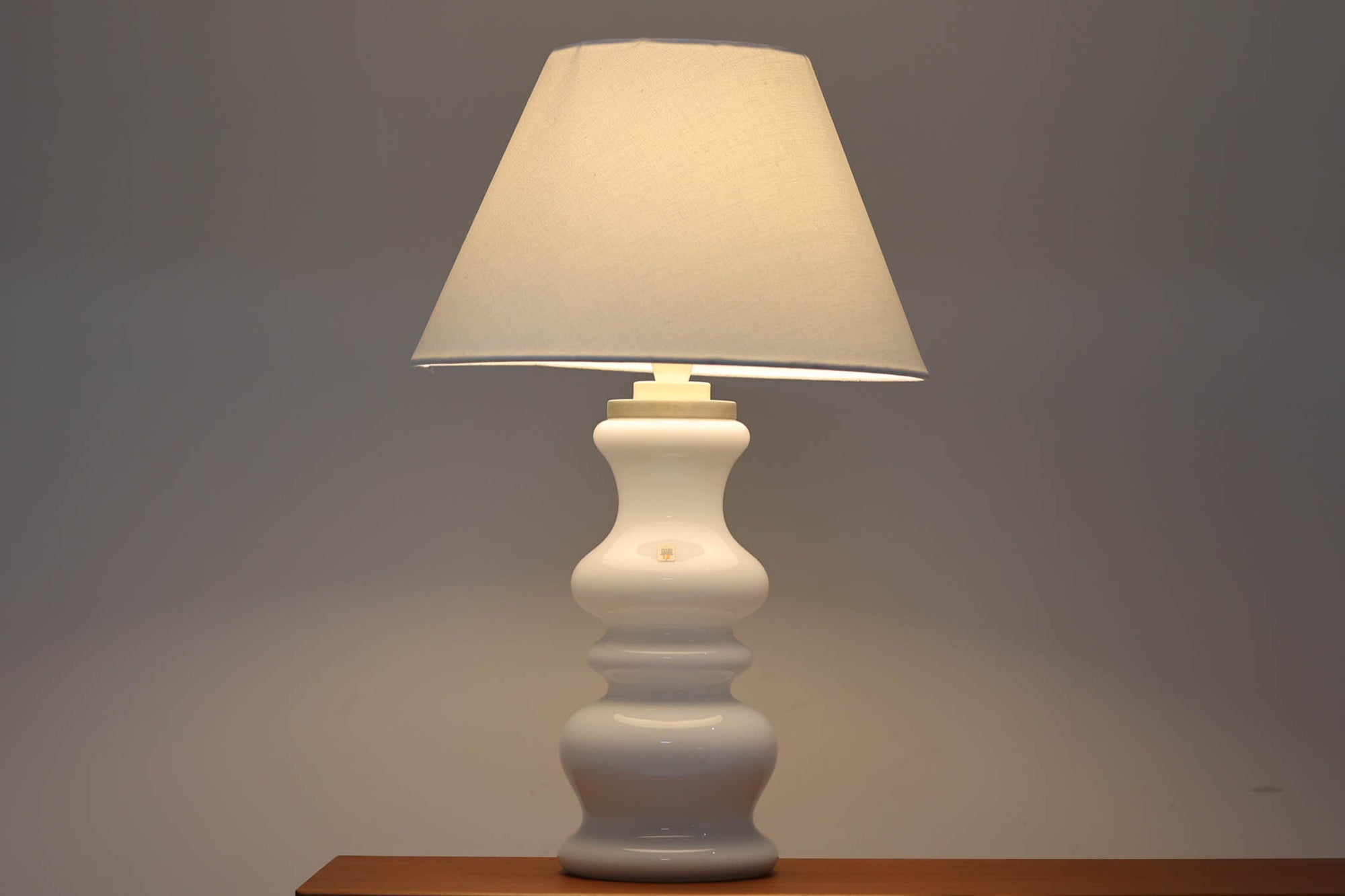 著名なデザイナー、Sidse Werner（シセ・ヴェルナー）による「BIBLIOTEK」のテーブルランプです。ペンダントランプはよく見かけますが、テーブルランプは非常に珍しいモデルです。ホルムガードならではの厚みのある高品質なガラスを使用しており、点灯するとシェードを通して柔らかな温かみのある光が広がり、空間を優雅に照らします。高さ64cmの大型サイズで存在感があり、リビングや書斎、ベッドサイドなど、あらゆるインテリアに美しく調和します。