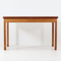 Ejner Larsen & Aksel Bender Madsenが手掛け、Naestved Mobelfabrikで製作されたエクステンションダイニングテーブルは、チーク材の天板とオーク材の脚部を組み合わせた北欧ビンテージらしい上質な佇まいが魅力です。直線的で端正なフォルムの中に、天板縁の丁寧な面取りや幕板の滑らかな造形など、当時の高い職人技が随所に感じられます。幅120cmから最大230cmまで伸長可能な構造により、日常使いから来客時まで柔軟に対応でき、空間や人数に合わせた使い方が可能です。経年変化によって深みを増した木肌の表情が、長く愛用されてきたビンテージならではの存在感を放ち、ダイニングの主役として落ち着きと品格を添えてくれる一台です。脚先には床へのダメージや汚れを防ぐためにチーク材がジョイントされており、実用性を考慮した当時の設計思想がうかがえます。また、裏面には「SKM」の刻印が確認でき、スウェーデン軍（Svenska KrigsMakten）の備品として使用されていたことを示す、背景のある希少な個体です。