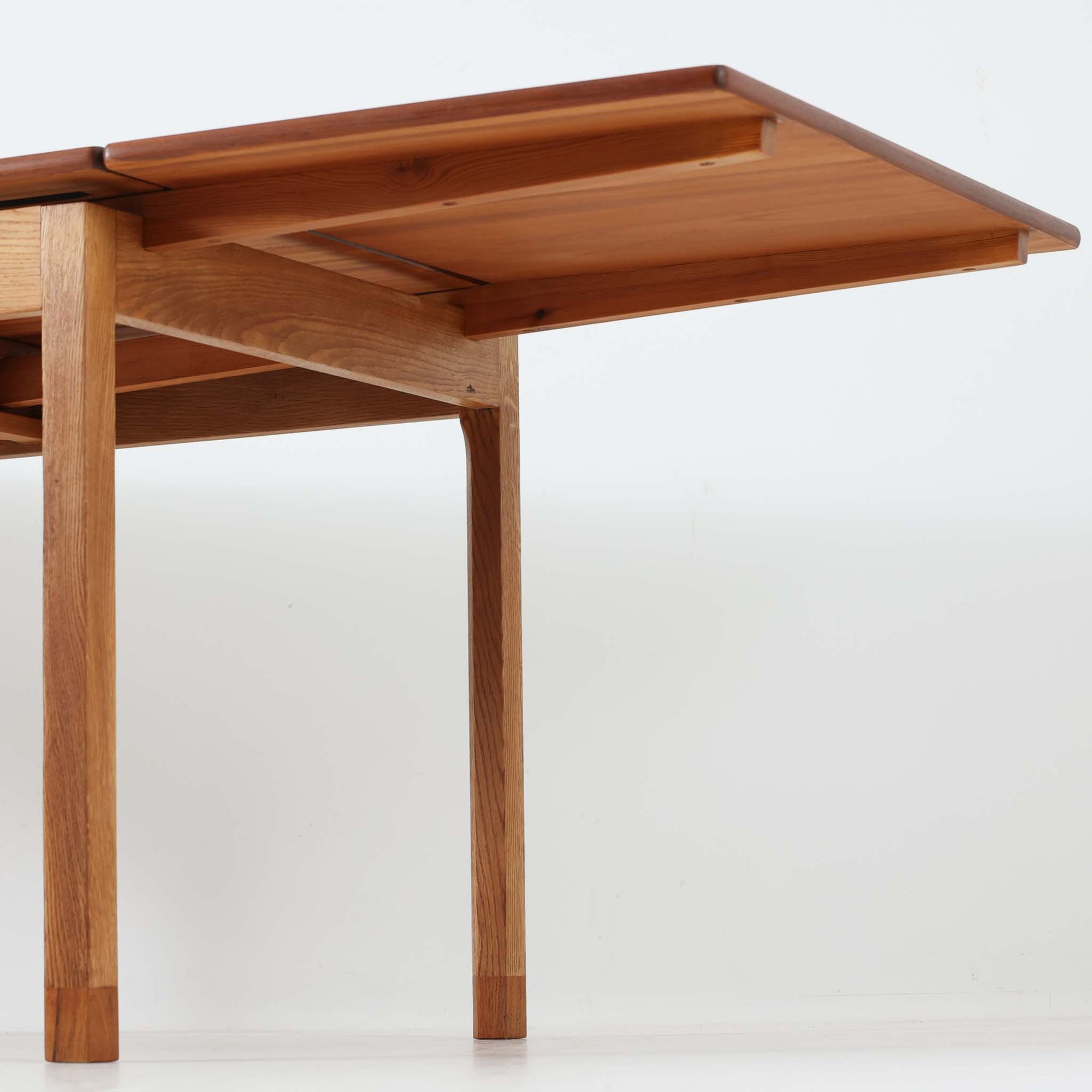 Ejner Larsen &amp; Aksel Bender Madsenが手掛け、Naestved Mobelfabrikで製作されたエクステンションダイニングテーブルは、チーク材の天板とオーク材の脚部を組み合わせた北欧ビンテージらしい上質な佇まいが魅力です。直線的で端正なフォルムの中に、天板縁の丁寧な面取りや幕板の滑らかな造形など、当時の高い職人技が随所に感じられます。幅120cmから最大230cmまで伸長可能な構造により、日常使いから来客時まで柔軟に対応でき、空間や人数に合わせた使い方が可能です。経年変化によって深みを増した木肌の表情が、長く愛用されてきたビンテージならではの存在感を放ち、ダイニングの主役として落ち着きと品格を添えてくれる一台です。脚先には床へのダメージや汚れを防ぐためにチーク材がジョイントされており、実用性を考慮した当時の設計思想がうかがえます。また、裏面には「SKM」の刻印が確認でき、スウェーデン軍（Svenska KrigsMakten）の備品として使用されていたことを示す、背景のある希少な個体です。