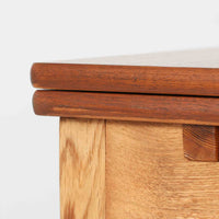 Ejner Larsen & Aksel Bender Madsenが手掛け、Naestved Mobelfabrikで製作されたエクステンションダイニングテーブルは、チーク材の天板とオーク材の脚部を組み合わせた北欧ビンテージらしい上質な佇まいが魅力です。直線的で端正なフォルムの中に、天板縁の丁寧な面取りや幕板の滑らかな造形など、当時の高い職人技が随所に感じられます。幅120cmから最大230cmまで伸長可能な構造により、日常使いから来客時まで柔軟に対応でき、空間や人数に合わせた使い方が可能です。経年変化によって深みを増した木肌の表情が、長く愛用されてきたビンテージならではの存在感を放ち、ダイニングの主役として落ち着きと品格を添えてくれる一台です。脚先には床へのダメージや汚れを防ぐためにチーク材がジョイントされており、実用性を考慮した当時の設計思想がうかがえます。また、裏面には「SKM」の刻印が確認でき、スウェーデン軍（Svenska KrigsMakten）の備品として使用されていたことを示す、背景のある希少な個体です。