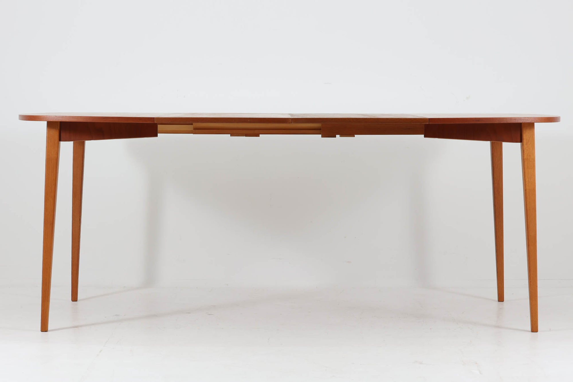 こちらはスウェーデンで買い付けたエクステンションダイニングテーブルです。天板は、珍しいラウンドスクエアの形状を採用しており、柔らかな印象と洗練されたデザインが魅力です。脚部は左右に開く構造で安定感があり、幕板は中央部分に向かってカットされているため、椅子の出し入れや座り心地にも配慮されています。また、来客時には付属の拡張板を使用することで、天板を簡単に広げられ、大人数での食事にも対応可能です。使い手の利便性を追求した設計が光る一品です。素材には、良質なチーク材とオーク材を使用しており、木目の美しさや温かみを感じられます。北欧家具ならではの高いデザイン性と実用性を兼ね備えたダイニングテーブルをぜひお楽しみください。