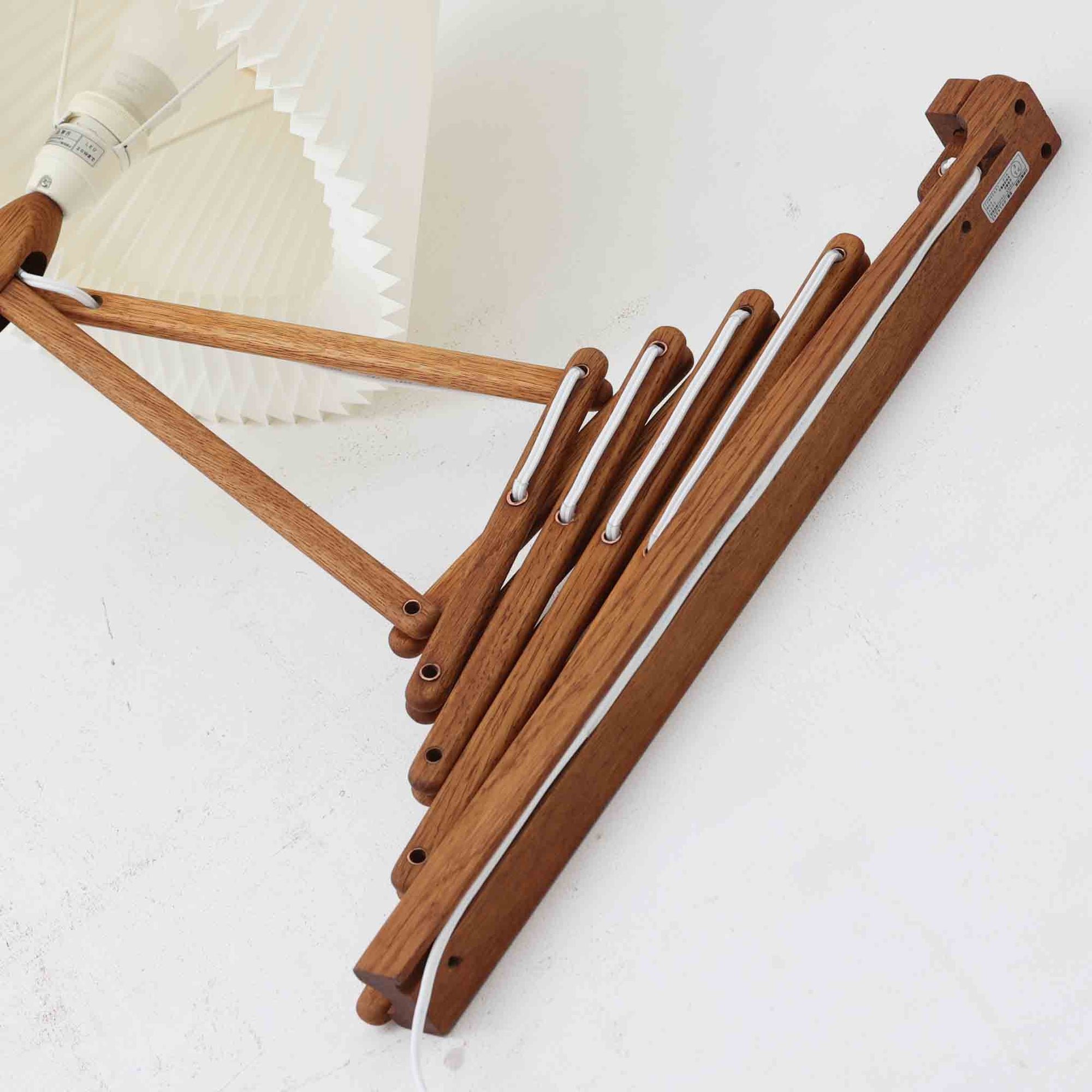 Erik Hansen（エリック・ハンセン）がデザインし、Le Klint（レ・クリント）から発表されたブラケットランプModel 332です。こちらは現行品には見られない一回り大きなサイズ感が特徴で、存在感のある佇まいを持っています。オーク材のナチュラルな風合いと、職人によるプリーツシェードが織りなす柔らかな光が空間を優しく包み込みます。アコーディオン式のアームは伸縮可能で、ベッドサイドやリビングの読書灯としても活躍する実用性の高い一品です。