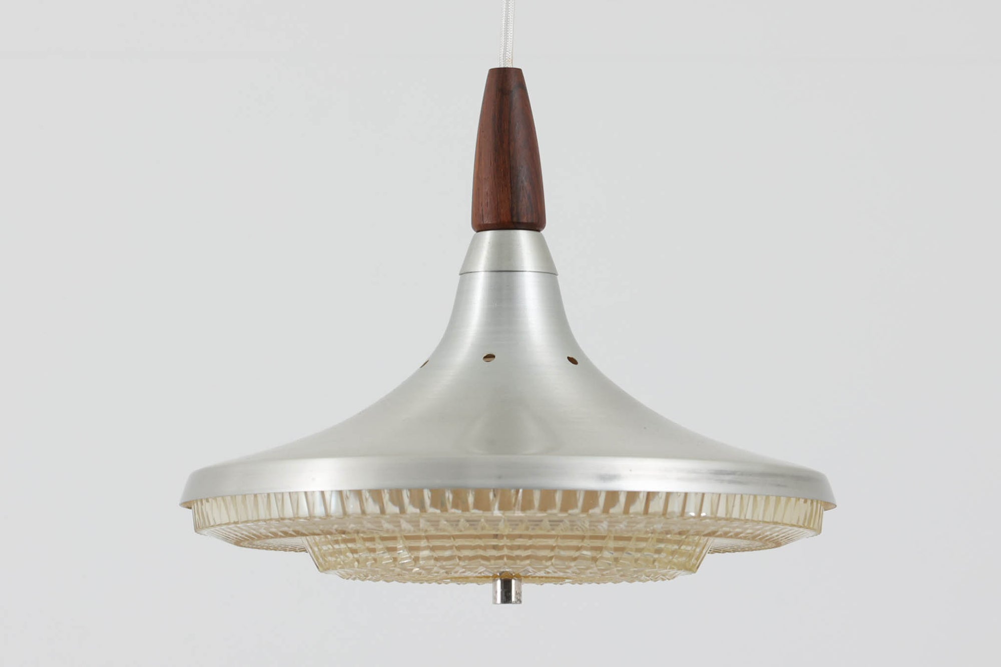 北欧より買い付けたこのペンダントライトは、絞り出された形状のアルミと、トップにジョイントされたウッドのコントラストが美しい一品です。ボトムカバーには装飾されたプラスチックが使用され、シェードの上部には細やかな穴が開けられています。その穴とボトムカバーからこぼれ出る光が、空間に柔らかな美しさを与え、独特の光の漏れ具合が洗練された雰囲気を演出します。