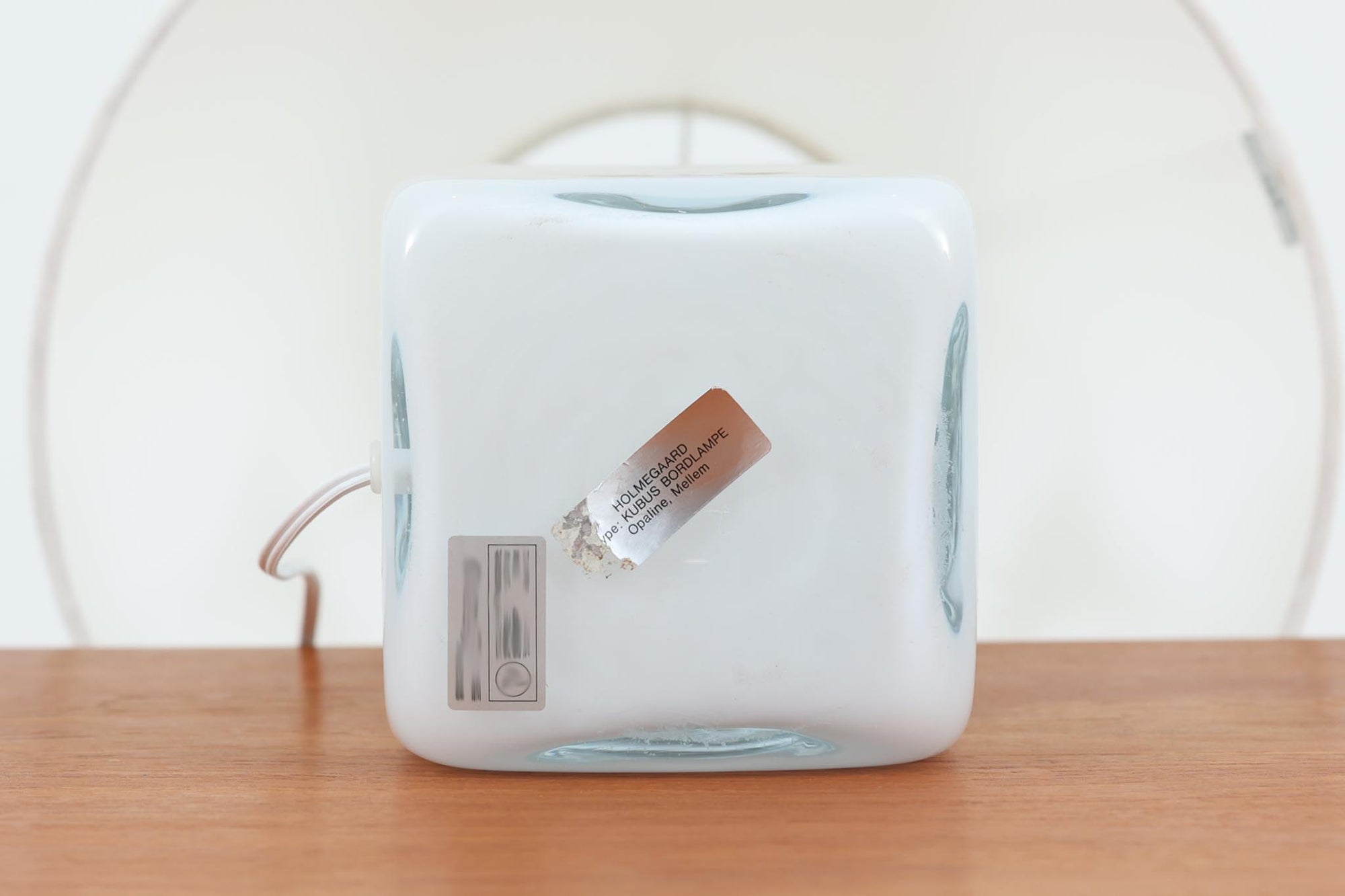 Per Lütken（ペル・ルッケン）が1980年代にデザインした「Kubus（キューブス）」シリーズのテーブルランプです。名前の通り、立方体（キューブ）をモチーフにしたホルムガードならではの厚みのある高品質なガラスが使用されており、点灯すると柔らかな光が広がり、空間に温かみと落ち着きを与えます。細いメタルの支柱が全体を引き締め、ミニマルで洗練されたデザインを際立たせています。大中小のサイズ展開がある中で、こちらは中サイズのモデルです。