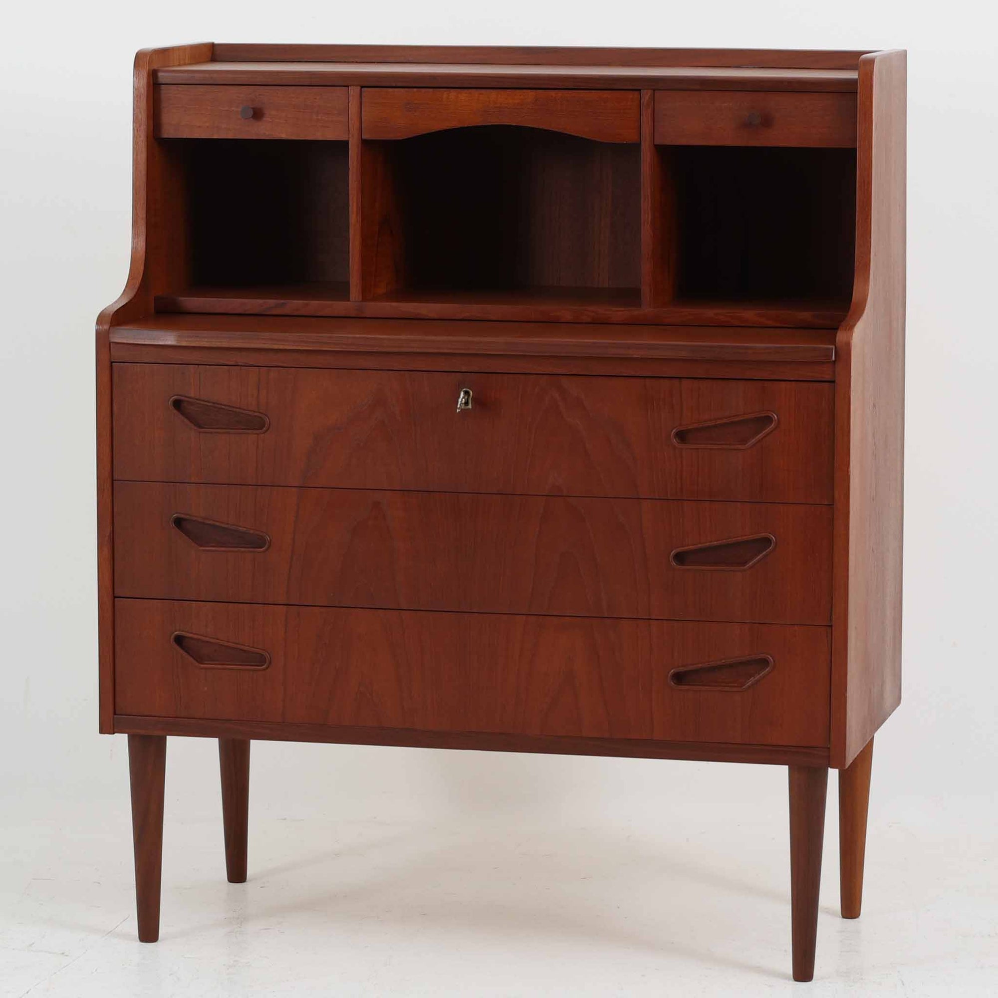 デンマーク製ライティングビューロです。上段には小引き出しと仕切り棚、中央には小さなミラーが備わり、書き物や身支度など多用途にお使いいただけます。チーク材の深みある色合いと滑らかな木目が美しく、シンプルで直線的なフォルムが落ち着いた印象を与えます。引き出しの斜めに削り出された取っ手やテーパー脚など、北欧らしい洗練されたディテールが魅力です。経年により増した艶が味わい深く、リビングや寝室に上品な温もりを添える一台です。