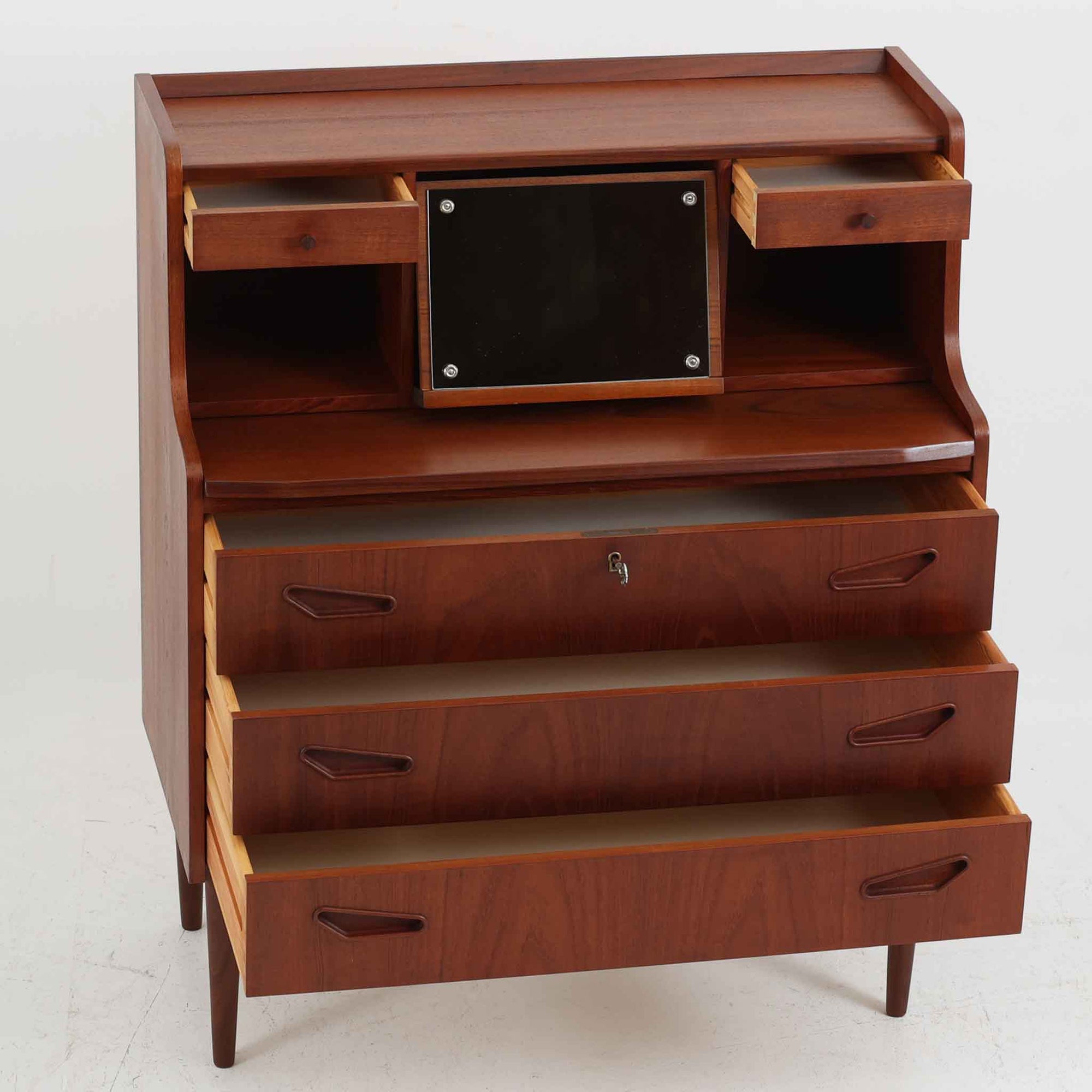 デンマーク製ライティングビューロです。上段には小引き出しと仕切り棚、中央には小さなミラーが備わり、書き物や身支度など多用途にお使いいただけます。チーク材の深みある色合いと滑らかな木目が美しく、シンプルで直線的なフォルムが落ち着いた印象を与えます。引き出しの斜めに削り出された取っ手やテーパー脚など、北欧らしい洗練されたディテールが魅力です。経年により増した艶が味わい深く、リビングや寝室に上品な温もりを添える一台です。