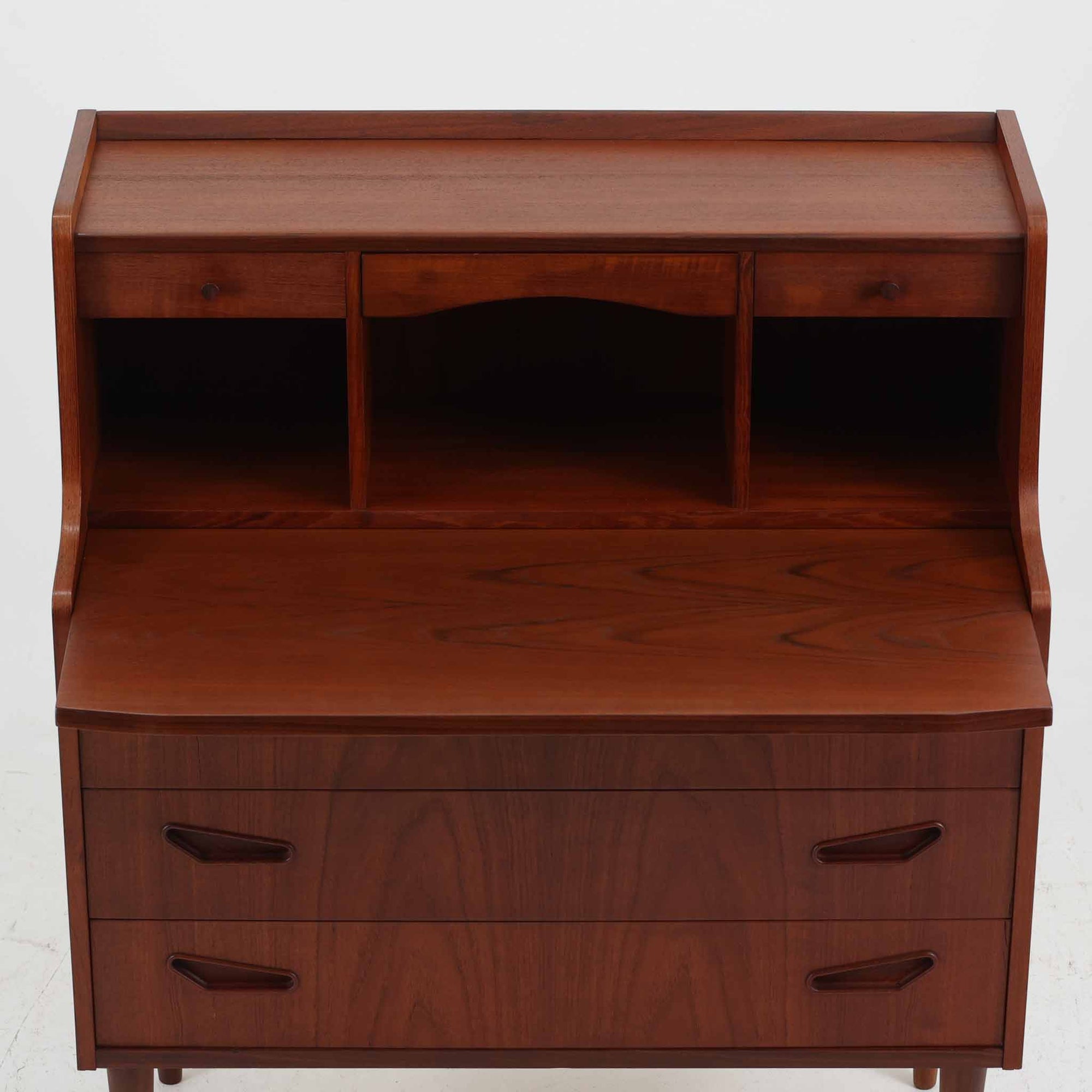 デンマーク製ライティングビューロです。上段には小引き出しと仕切り棚、中央には小さなミラーが備わり、書き物や身支度など多用途にお使いいただけます。チーク材の深みある色合いと滑らかな木目が美しく、シンプルで直線的なフォルムが落ち着いた印象を与えます。引き出しの斜めに削り出された取っ手やテーパー脚など、北欧らしい洗練されたディテールが魅力です。経年により増した艶が味わい深く、リビングや寝室に上品な温もりを添える一台です。