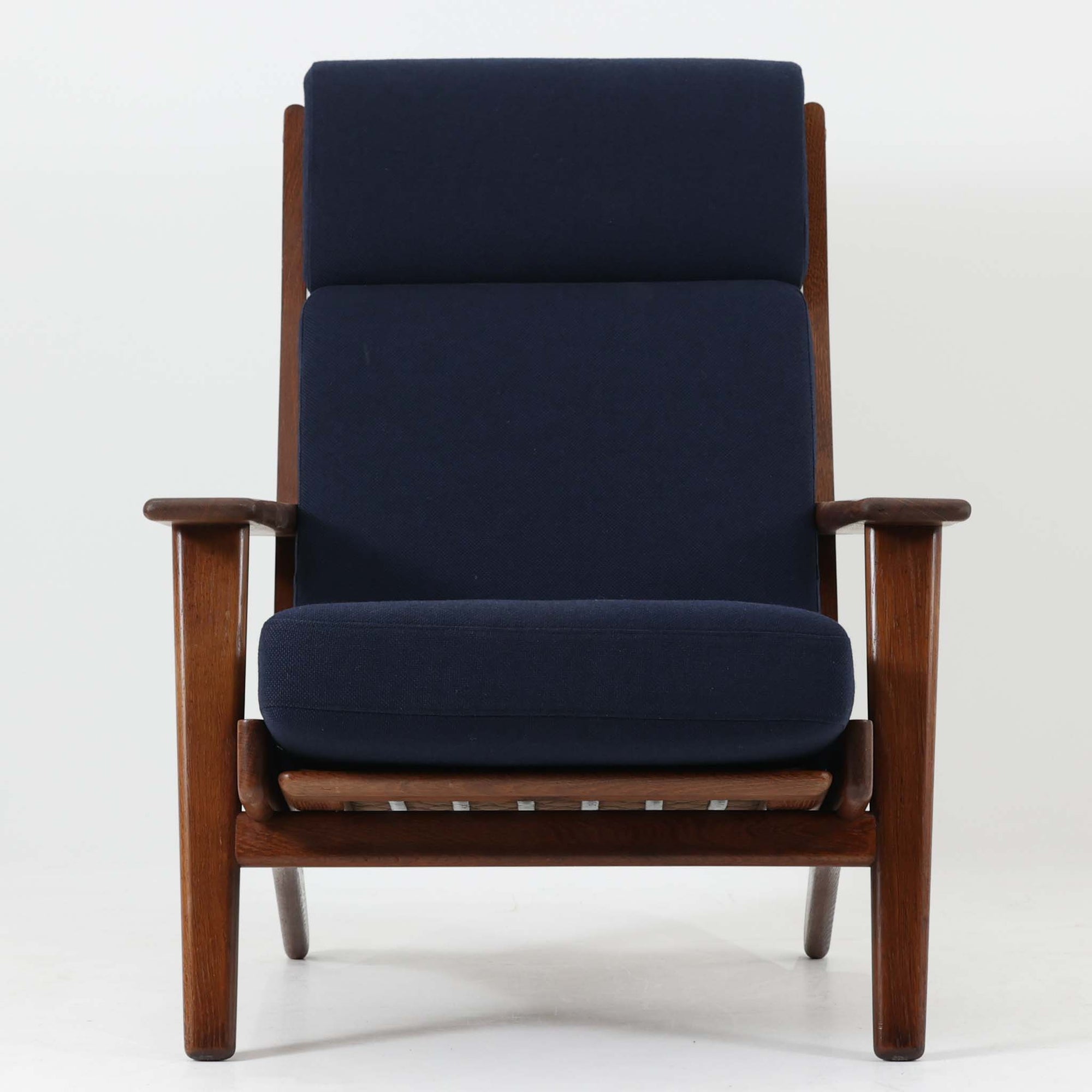 Hans J. Wegnerがデザインし、Getamaが製作したGE290Aハイバックシングルソファです。ダークオーク材の堅牢なフレームは丸みを帯びたアームと後方へ傾斜した脚部が特徴で、身体をやさしく受け止める構造美が際立ちます。張り替えられた落ち着いた色調のクッションは、内部にビンテージ特有のスプリングを内包し、程よい弾力と快適な座り心地を実現。読書や寛ぎの時間に最適で、経年により深みを増した木部の質感が北欧ビンテージならではの存在感と上質な雰囲気を空間に添える一脚です。