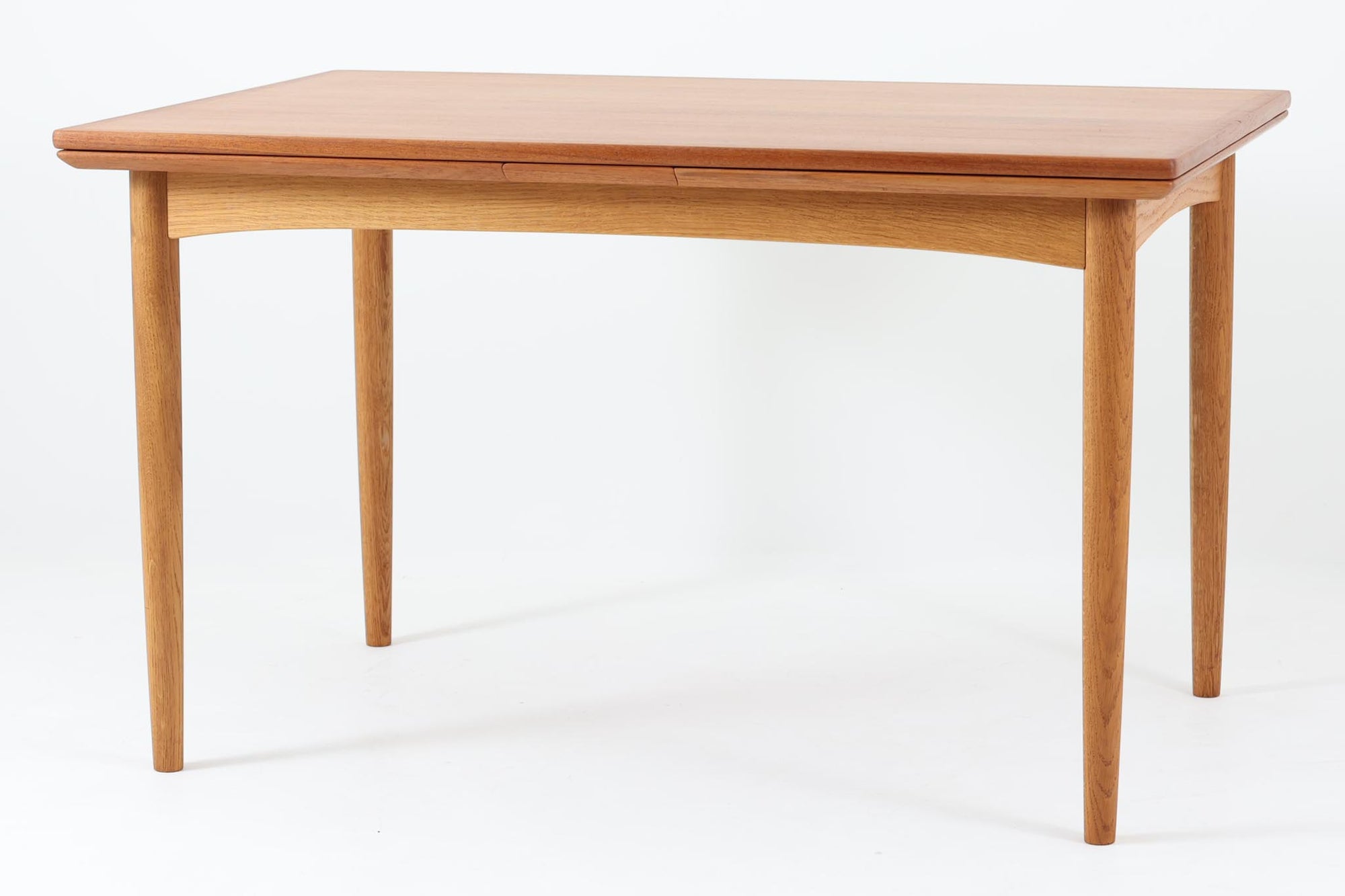 デンマークの名工Slagelse mobelvaerk社が手掛けたこちらのダイニングテーブルは、アーチ状の幕板が特徴であり、異なる素材の組み合わせが美しさを際立たせます。天板には上質なチーク材が用いられ、脚部にはナチュラルなオーク材が採用されています。また、幕板は強度と使い勝手が考慮され、通常時には4人、拡張時には6～8人が快適に使用できる設計となっています。