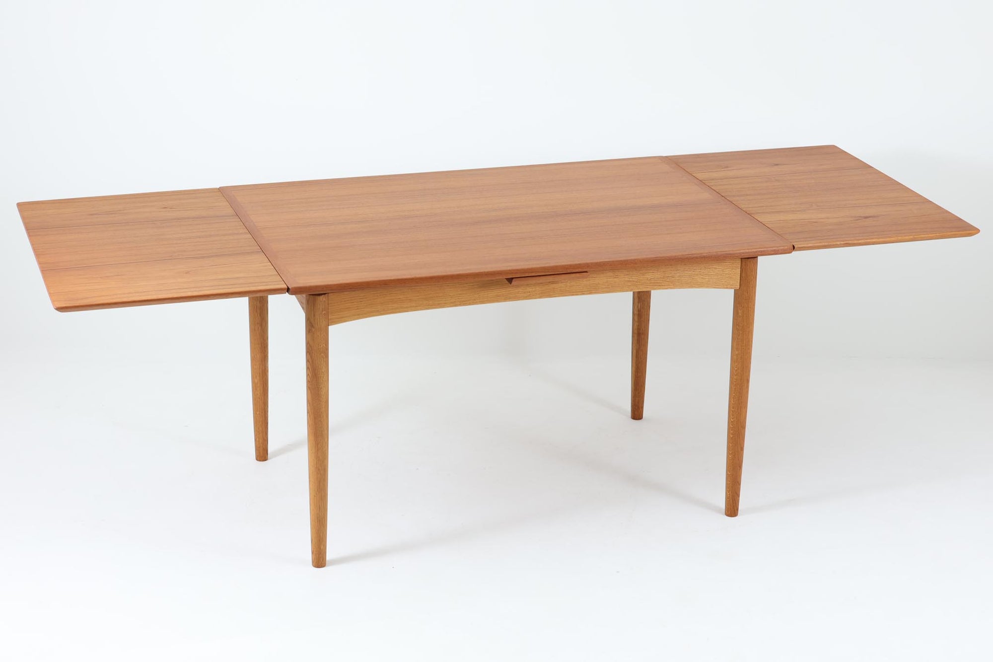 デンマークの名工Slagelse mobelvaerk社が手掛けたこちらのダイニングテーブルは、アーチ状の幕板が特徴であり、異なる素材の組み合わせが美しさを際立たせます。天板には上質なチーク材が用いられ、脚部にはナチュラルなオーク材が採用されています。また、幕板は強度と使い勝手が考慮され、通常時には4人、拡張時には6～8人が快適に使用できる設計となっています。