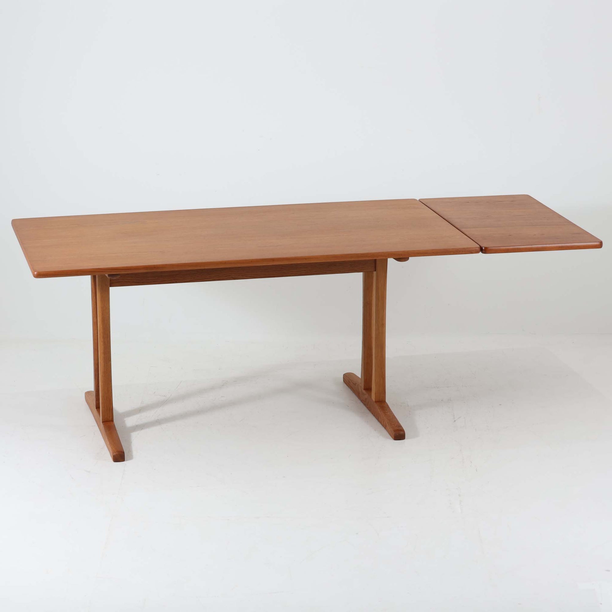 Borge Mogensen（ボーエ・モーエンセン）がFDB Moblerのために手掛けたC18ダイニングテーブルは、チーク材天板とオーク材脚部を組み合わせた北欧モダンを象徴するビンテージ家具で、直線的で端正なプロポーションと安定感のあるT字脚構造が特徴です。拡張板を備え、来客時や家族構成の変化にも柔軟に対応でき、日常使いから特別な食卓まで幅広く活躍します。現在はCarl Hansen &amp; Son社より復刻生産されていますが、チーク材天板とオーク材脚部という素材の組み合わせは当時のビンテージ品ならではの仕様で、脚部に用いられた真鍮製の留め具も現行品とは異なる形状となっています。経年により深みを増した木肌と、実用性を重視したモーエンセンらしい設計思想、そしてビンテージならではのディテールを味わえる一台です。