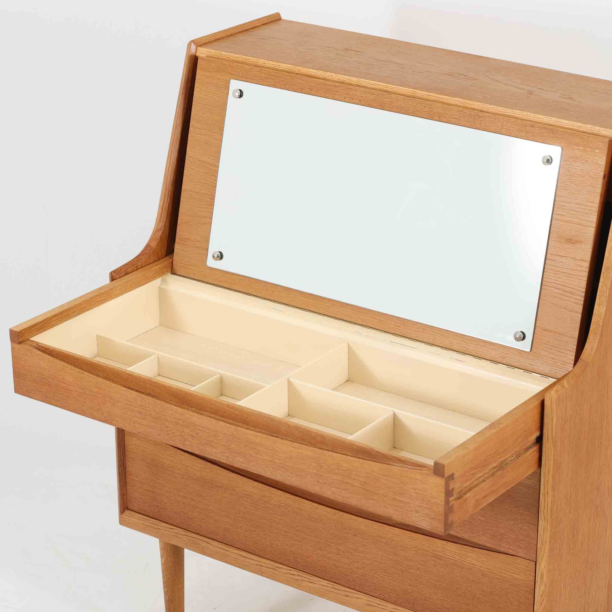 Arne Vodderがデザインし、Sibast Mobelfabrikが製造したオーク材のチェストドレッサーです。上部には小引出しや書類用の仕切りを備え、手前の天板を開くとミラーと細かな収納スペースが現れる機能的な構造で、下部の大容量引出しと合わせて身支度用品を美しく整理できます。弧を描く引出し前板や先細りの脚部など、職人技を感じさせる優雅な造形も魅力で、経年によって深まった木肌が空間に温かな存在感を添える希少な一台です。