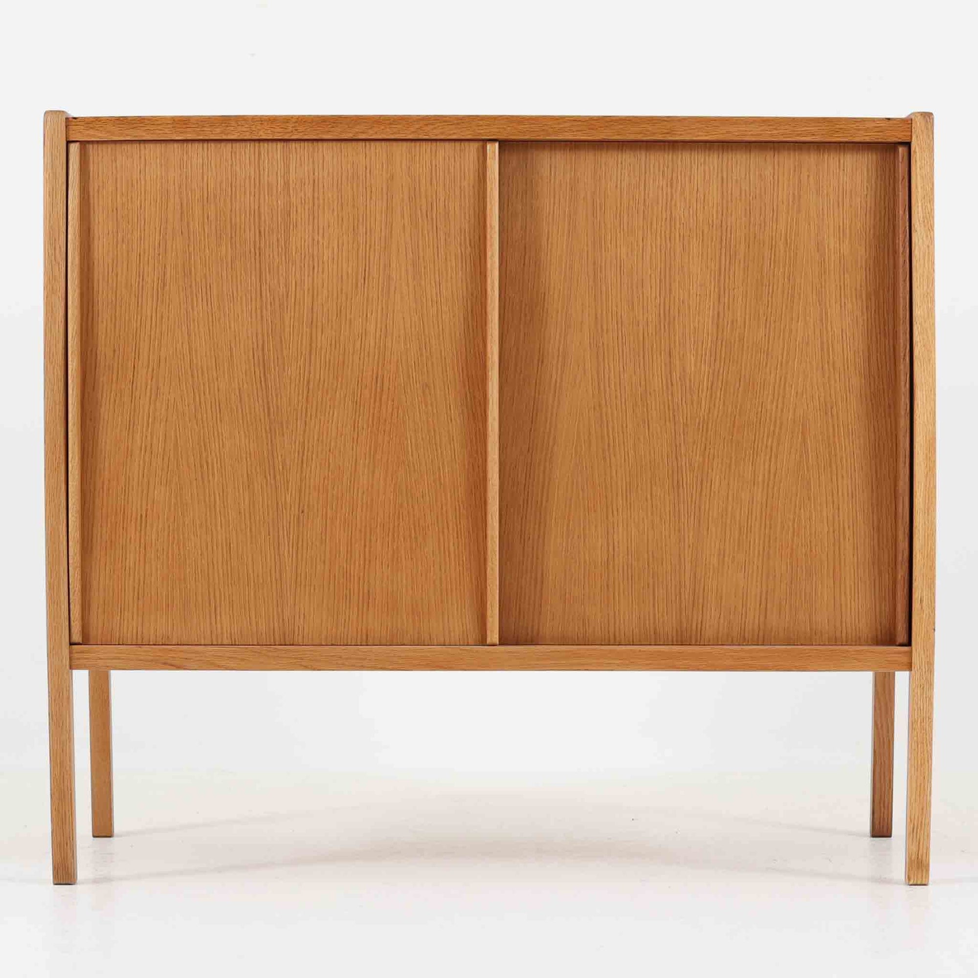 B. Fridhagenがデザインし、スウェーデンのBodafors社によって製造されたこちらのボードは、上質なオーク材を用いた小ぶりなサイズ感と、無駄を省いた洗練された意匠が魅力の一台です。スライド扉を開けると左右に仕切られた収納部が現れ、日用品や書籍の整理に最適です。細身の脚と角の立ったフレームが全体に軽やかな印象を与え、空間を圧迫せず設置できます。シンプルながらも素材の美しさと北欧らしい実用性が際立つ、希少なビンテージ家具です。