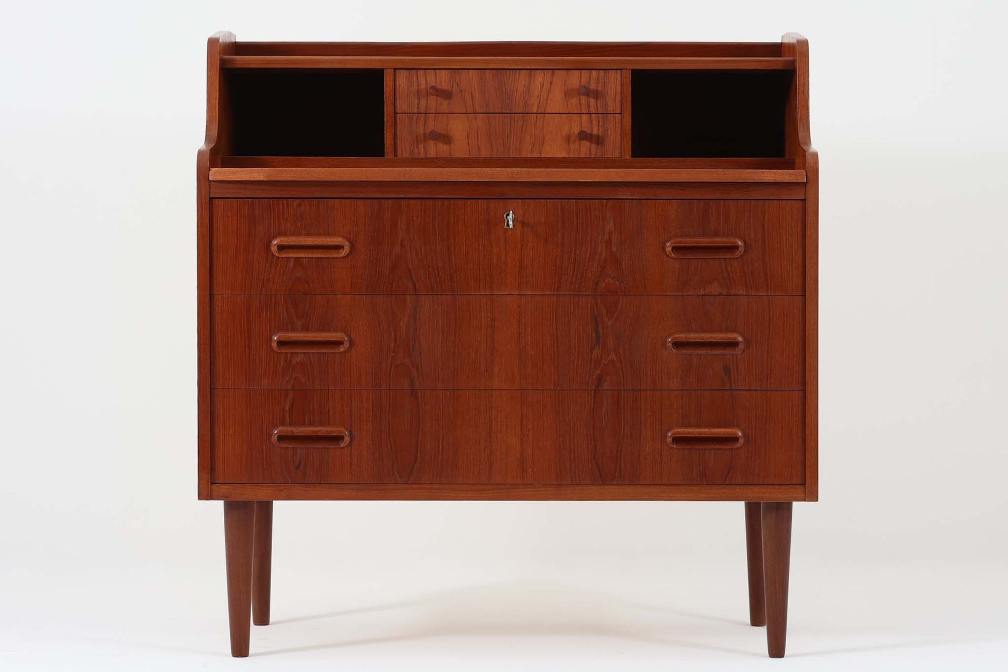 デンマークから買い付けたヴィンテージのライティングビューロです。高さは88cmと小ぶりで、コンパクトなサイズ感が魅力です。上段には2杯の小引き出しと収納スペースが備わっており、細々としたアイテムを整理して収納できます。下段には3杯の大きな引き出しがあり、1段目には鍵が付属しているため、貴重品の保管にも最適です。また、天板を引き出すことでデスクとしてもお使いいただけるため、簡単な作業や書き物にも対応。普段はチェストとして使用したり、雑貨をディスプレイしたりと、幅広い用途で活躍します。本体には良質なチーク材が使用されており、美しい木目が北欧家具ならではの上品さを引き立てています。