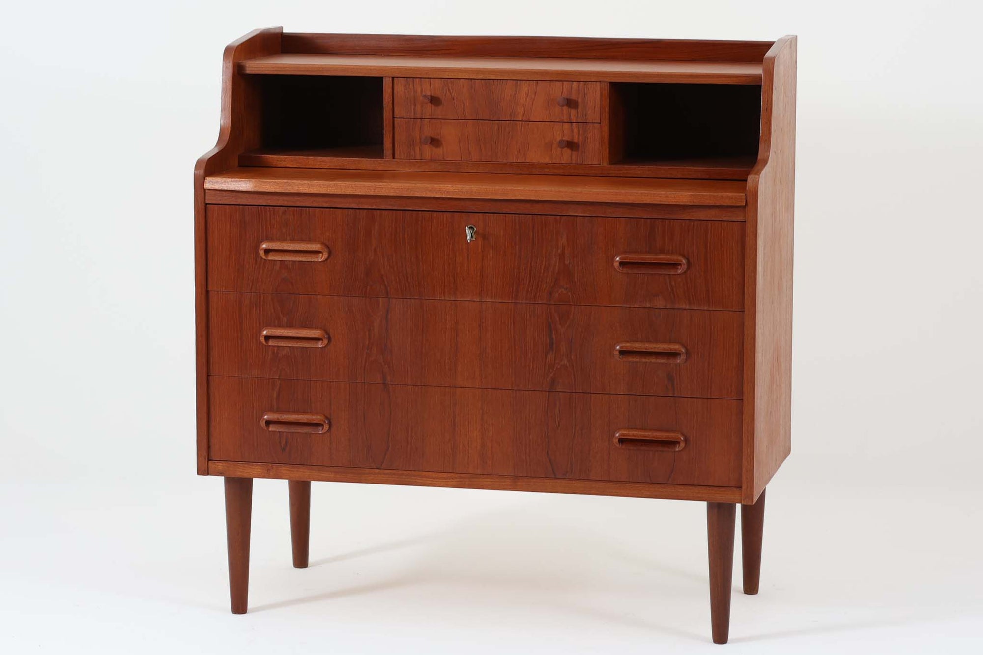 デンマークから買い付けたヴィンテージのライティングビューロです。高さは88cmと小ぶりで、コンパクトなサイズ感が魅力です。上段には2杯の小引き出しと収納スペースが備わっており、細々としたアイテムを整理して収納できます。下段には3杯の大きな引き出しがあり、1段目には鍵が付属しているため、貴重品の保管にも最適です。また、天板を引き出すことでデスクとしてもお使いいただけるため、簡単な作業や書き物にも対応。普段はチェストとして使用したり、雑貨をディスプレイしたりと、幅広い用途で活躍します。本体には良質なチーク材が使用されており、美しい木目が北欧家具ならではの上品さを引き立てています。