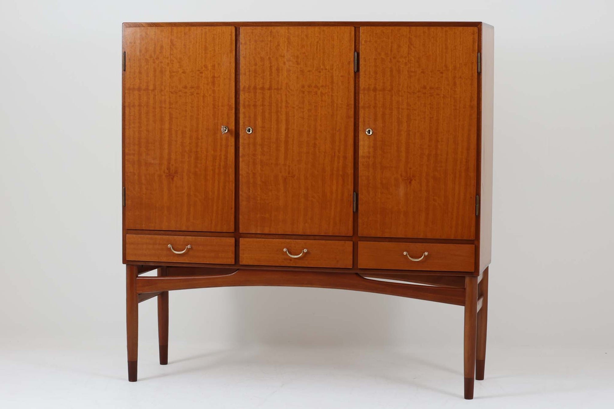 FDBモブラーでBorge mogensenの後任としてアートディレクターを務めたPoul m. Voltherによる、美しく洗練されたキャビネット。良質なマホガニー材の本体に、ビーチ材の脚部を組み合わせたコントラストが魅力です。真鍮の丸い鍵穴が特徴的な3枚の扉を備え、中央には調節可能な棚、両側には精巧な蟻継ぎが施された移動式の引き出しを配しています。下部には真鍮の俵型ハンドル付きの引き出しが3杯並び、細部へのこだわりが際立ちます。また、室内で靴を履く文化に配慮し、脚先には汚れ防止のための濃い塗装が施されています。1950年代にデザインされた本作は、先細りの脚やストレッチャーがミニマルな美しさを際立たせる一方、シンメトリーなフォルムや真鍮の装飾が伝統的なエレガンスを醸し出し、モダンとクラシックが調和した逸品です。