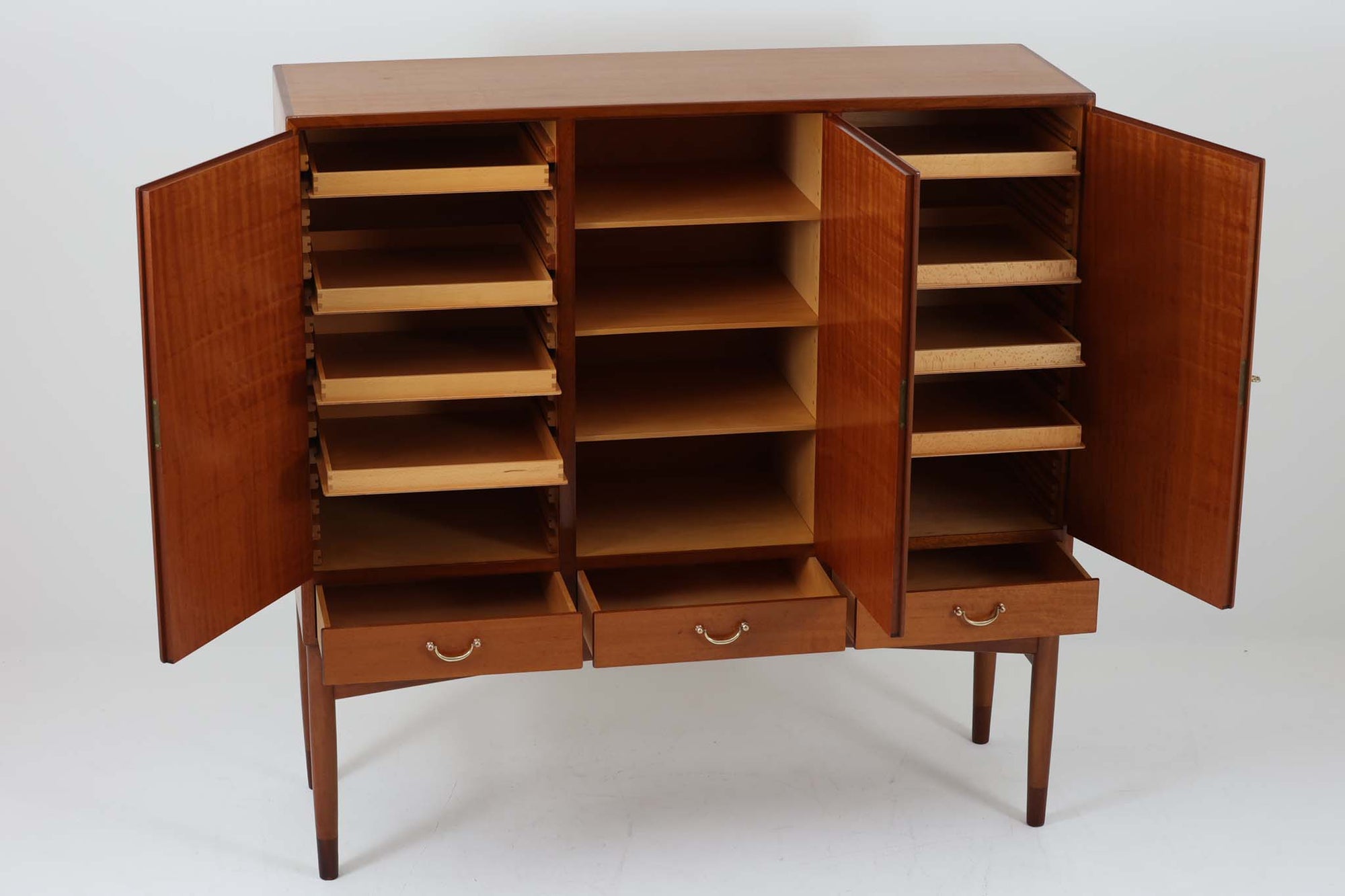 FDBモブラーでBorge mogensenの後任としてアートディレクターを務めたPoul m. Voltherによる、美しく洗練されたキャビネット。良質なマホガニー材の本体に、ビーチ材の脚部を組み合わせたコントラストが魅力です。真鍮の丸い鍵穴が特徴的な3枚の扉を備え、中央には調節可能な棚、両側には精巧な蟻継ぎが施された移動式の引き出しを配しています。下部には真鍮の俵型ハンドル付きの引き出しが3杯並び、細部へのこだわりが際立ちます。また、室内で靴を履く文化に配慮し、脚先には汚れ防止のための濃い塗装が施されています。1950年代にデザインされた本作は、先細りの脚やストレッチャーがミニマルな美しさを際立たせる一方、シンメトリーなフォルムや真鍮の装飾が伝統的なエレガンスを醸し出し、モダンとクラシックが調和した逸品です。