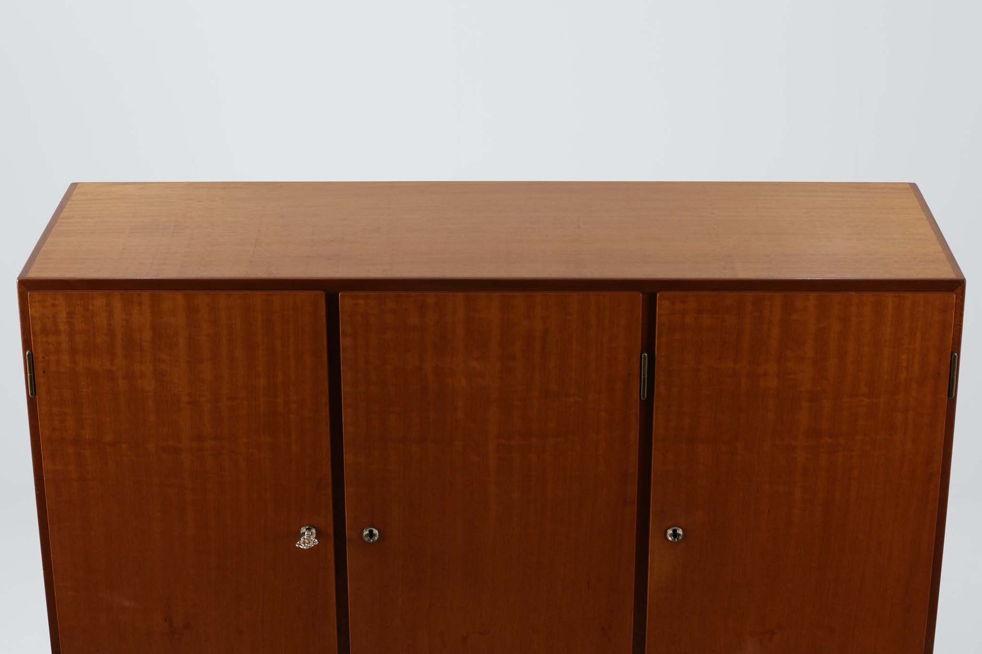 FDBモブラーでBorge mogensenの後任としてアートディレクターを務めたPoul m. Voltherによる、美しく洗練されたキャビネット。良質なマホガニー材の本体に、ビーチ材の脚部を組み合わせたコントラストが魅力です。真鍮の丸い鍵穴が特徴的な3枚の扉を備え、中央には調節可能な棚、両側には精巧な蟻継ぎが施された移動式の引き出しを配しています。下部には真鍮の俵型ハンドル付きの引き出しが3杯並び、細部へのこだわりが際立ちます。また、室内で靴を履く文化に配慮し、脚先には汚れ防止のための濃い塗装が施されています。1950年代にデザインされた本作は、先細りの脚やストレッチャーがミニマルな美しさを際立たせる一方、シンメトリーなフォルムや真鍮の装飾が伝統的なエレガンスを醸し出し、モダンとクラシックが調和した逸品です。