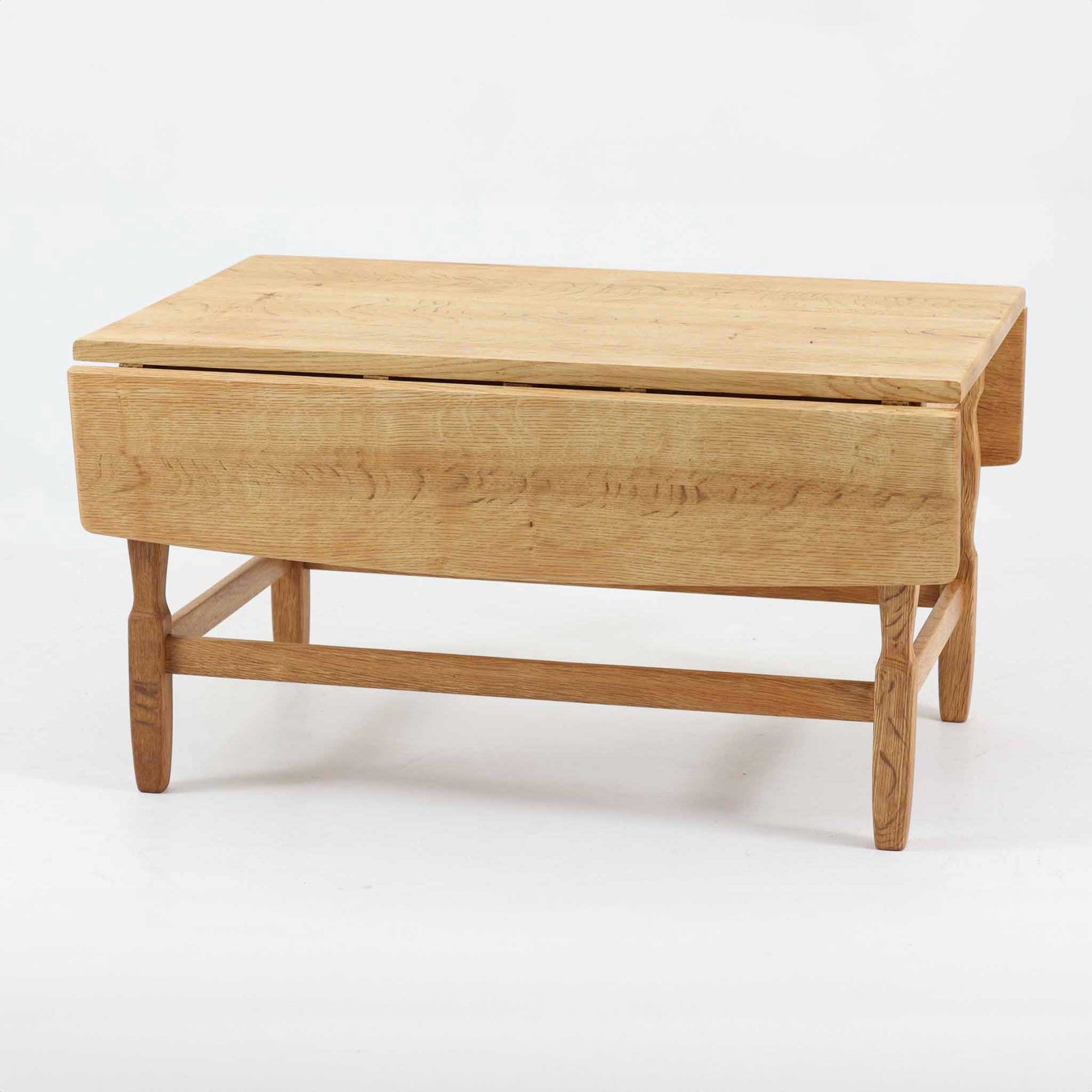 デンマークの建築家Henning Kjaernulf（ヘニング・ケアヌルフ）によってデザインされた、オーク無垢材のドロップリーフ式センターテーブルです。製造は高品質な家具で知られるEGモブラー社。無垢材ならではの美しい木目と、クラシカルで彫刻的な脚部のラインが印象的です。天板は両側に開閉式のリーフが備わっており、来客時や作業時には広げてスペースを拡張できるなど、実用性も兼ね備えています。テーブルの高さはやや高めの設計で、ソファ前に置いても手元が使いやすく、センターテーブルとして快適にお使いいただけます。必要に応じてサイドテーブルやティーテーブルとしても活躍する、用途の広い一台です。