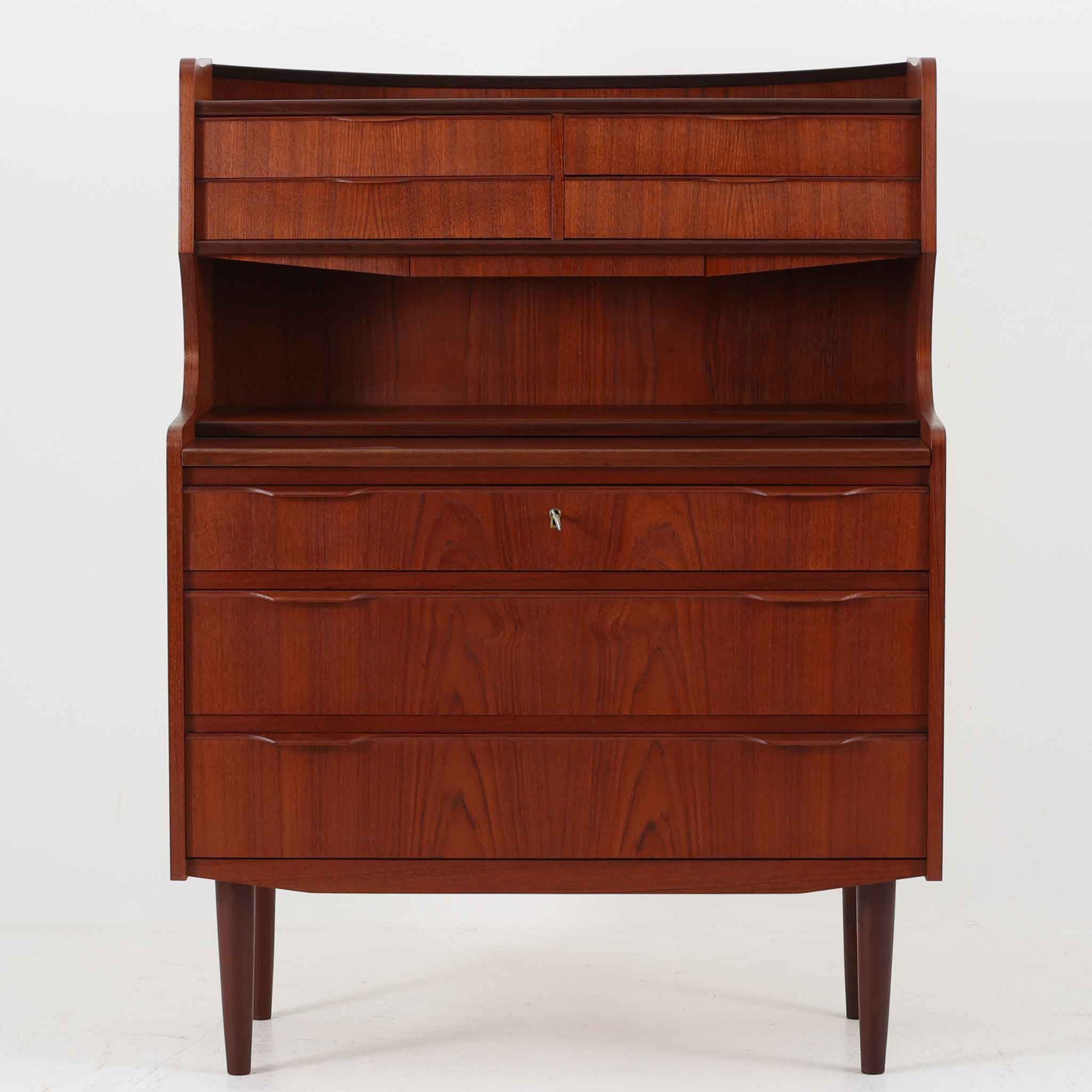 デンマーク製のチーク材によるライティングビューロです。幅85cmと程よいサイズ感で、上段には小引き出しを備え、小物や文具を整理しやすい構造になっています。中央のライティング天板は手前にスライドさせることで作業スペースが確保でき、書き物やノートPCの使用に最適です。さらに、内部には鏡が備わっており、ドレッサーとしても活用できる実用的なデザインです。下段には容量の大きな引き出しが3杯あり、衣類や書類の収納にも対応します。チーク材特有の温かみのある木目と経年による艶が味わい深く、実用性と美観を兼ね備えたビンテージ家具です。