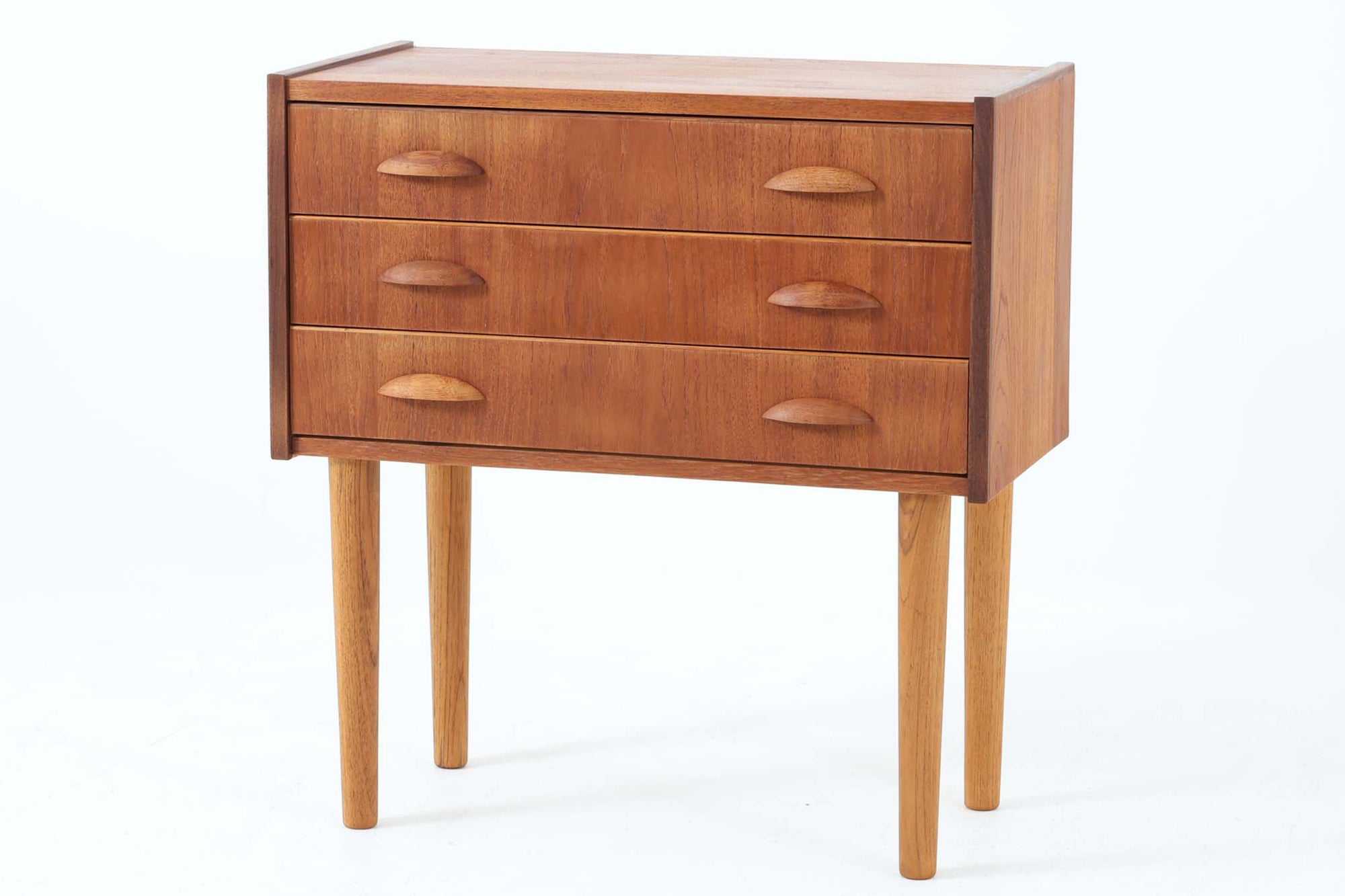 温かみのあるチーク材を用いた、デンマーク製のビンテージ3段チェストです。美しい木目と落ち着いた色合いが魅力で、引き出しには半月型の木製ハンドルがあしらわれています。直線的でシンプルなフォルムに、丸みを帯びたチーク材の脚が調和し、全体として統一感のある佇まいに仕上がっています。元の脚部に破損が見られたため、同じくチーク材で新たに作成しております。リビングや寝室、玄関など幅広い空間でお使いいただける実用的な収納家具です。