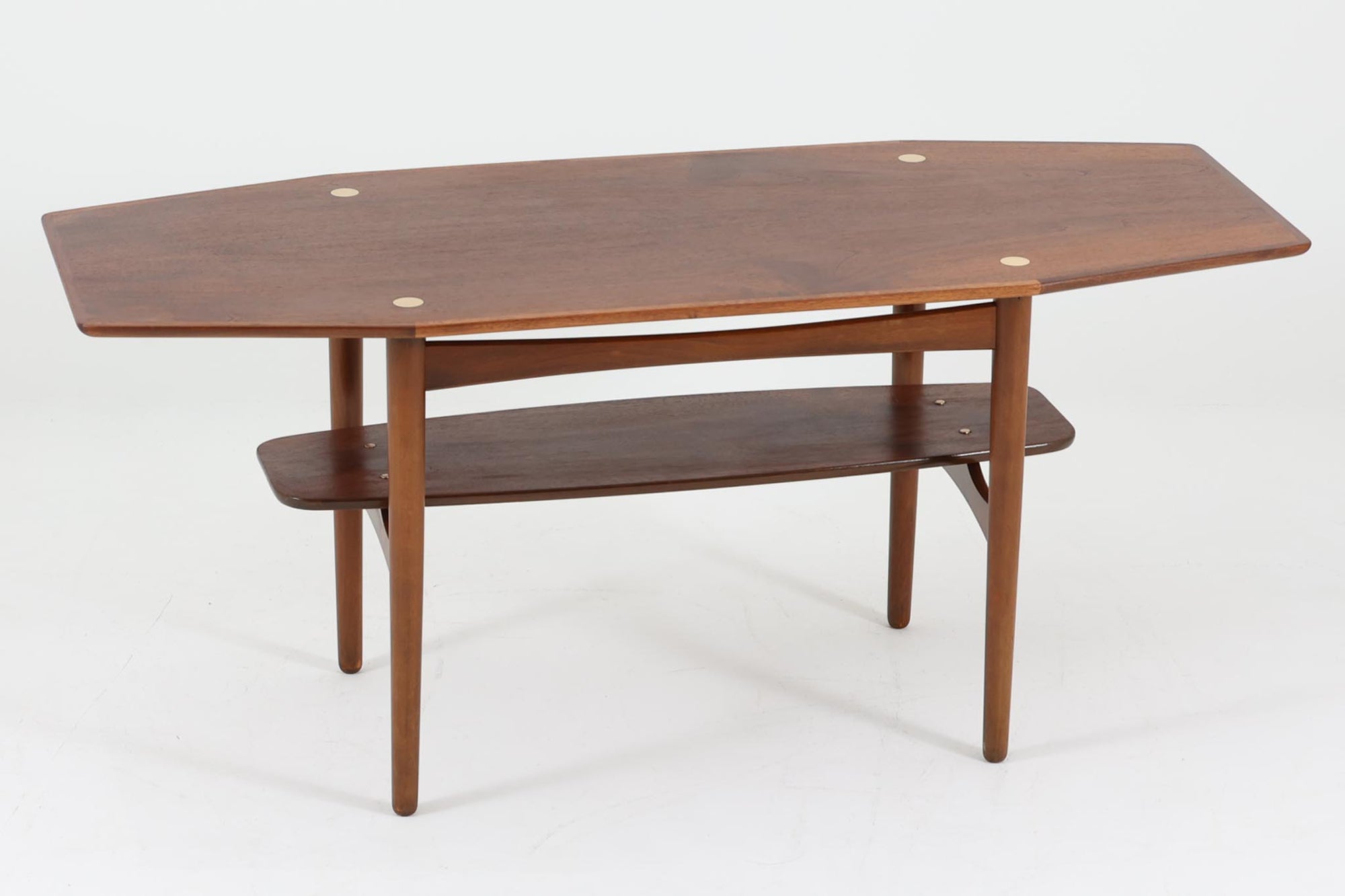 台形を基にした多角形の天板が特徴的な、美しいビンテージのセンターテーブル。心地よい木目が際立つウォールナット材の天板と、ナチュラルな風合いのビーチ材の組み合わせが、温かみのある佇まいを演出しています。天板と脚部、棚板の接続部には丸い真鍮があしらわれ、シンプルなフォルムの中に洗練されたアクセントを加えています。また、脚部をつなぐ幕板の形状も独特で、細部にまでこだわりが感じられるデザインです。デザイナーは不明ですが、細部の仕上げや素材選びから高いクオリティがうかがえる一品。モダンとクラシックが絶妙に調和した、上質なビンテージテーブルです。