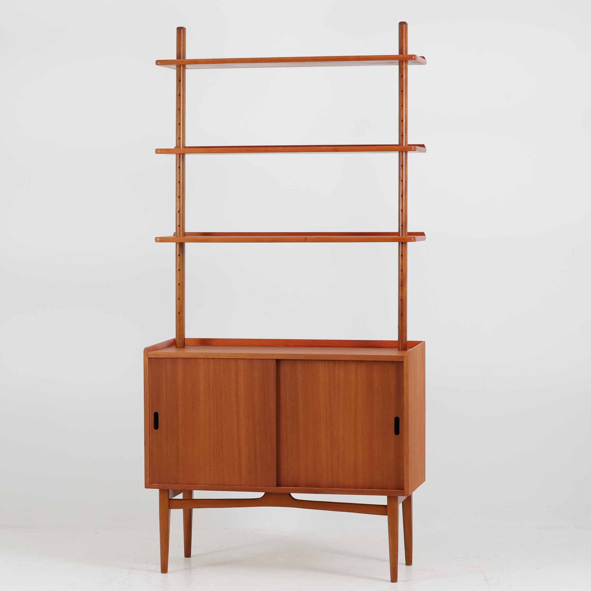 スウェーデンの老舗家具メーカー、Svenssons Mobler社によるマホガニー材のディスプレイシェルフです。上部の3段シェルフは高さを調整できる可動棚仕様で、書籍やオブジェ、植物などをバランス良く飾ることができます。下段のスライド扉収納は実用性が高く、生活感をすっきりと隠せる構造です。滑らかな木肌と温かみのある赤褐色の木目がマホガニーならではの上品な雰囲気を醸し出します。脚部の細身なテーパー形状も美しく、空間に軽やかさを添える北欧ビンテージ家具です。スライド扉の取っ手部分は丸くくり抜かれており、シンプルながらも洗練された印象を与えます。収納部と棚板の両サイドに開けられた穴に支柱を通して組み立てる独特の構造が特徴で、機能性とデザイン性を兼ね備えたスウェーデンらしい巧みな造りです。