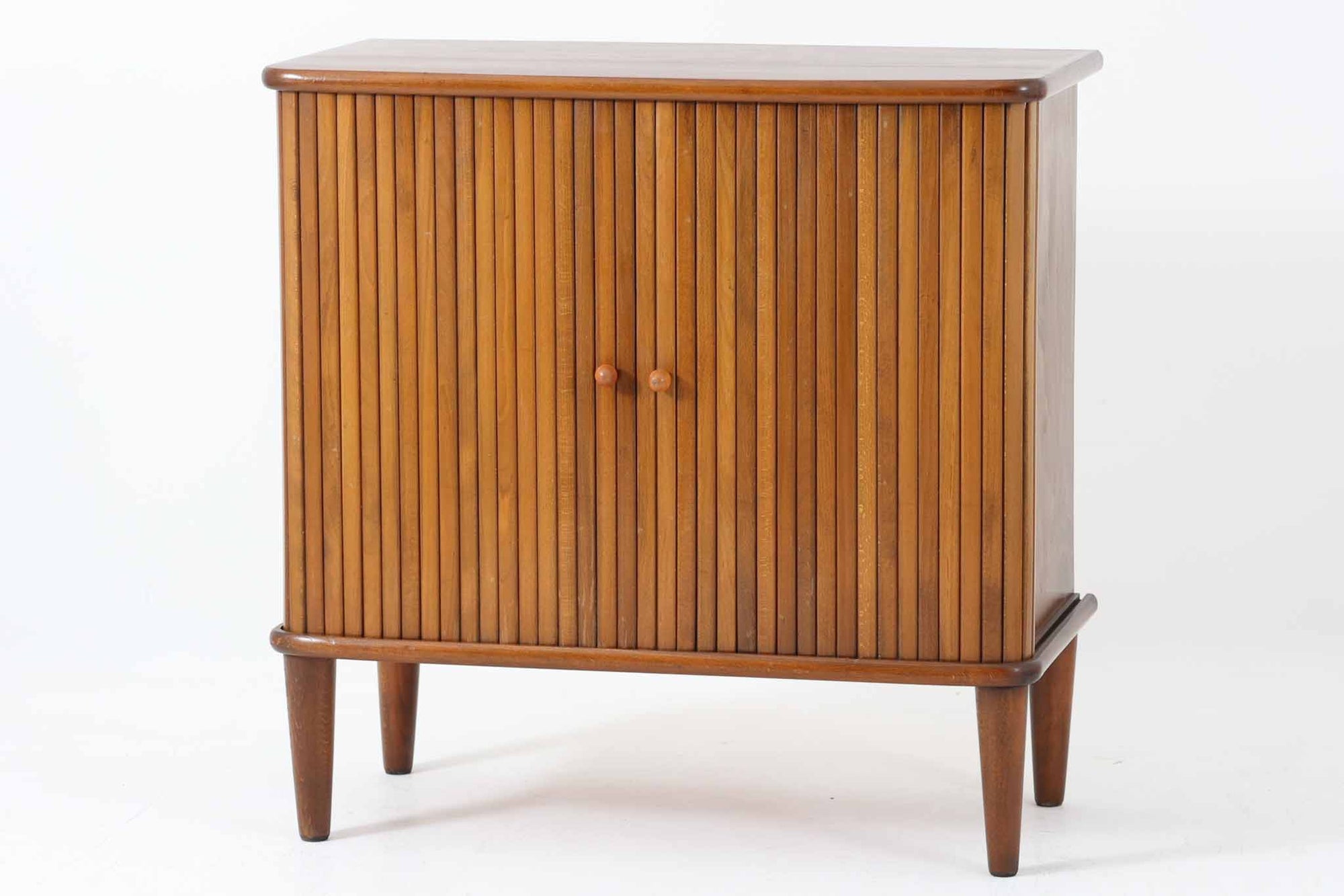 北欧デンマークで製造された、蛇腹扉が特徴的なビンテージキャビネットです。素材にはビーチ材と思われる無垢材や突板が使用されており、ステイン塗装により落ち着いたブラウンカラーに仕上げられています。前面には細かな縦ラインが連続する蛇腹扉が配され、開閉時に滑らかに動く美しい構造が魅力です。内部には棚板が一枚付属しており、収納としての機能性も十分です。丸脚や小さな丸い取っ手も北欧らしい柔らかなディテールで、リビングや玄関、寝室などさまざまな空間に自然に溶け込む一台です。