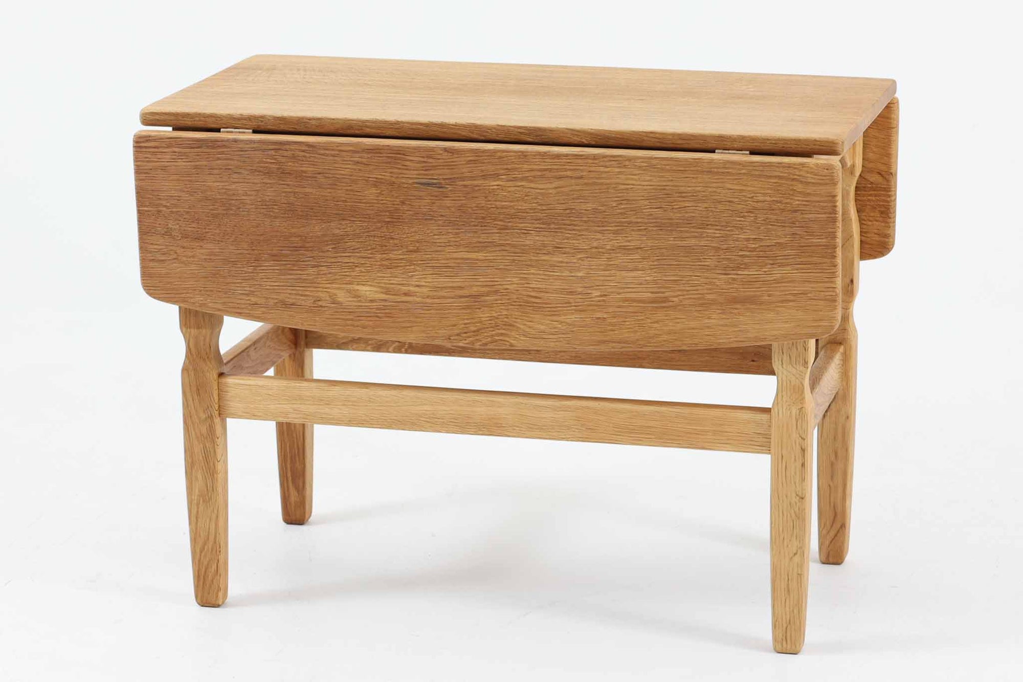 デンマークの建築家Henning Kjaernulf（ヘニング・ケアヌルフ）によってデザインされた、オーク無垢材のドロップリーフ式センターテーブルです。製造は高品質な家具で知られるEGモブラー社。無垢材ならではの美しい木目と、クラシカルで彫刻的な脚部のラインが印象的です。天板は両側に開閉式のリーフが備わっており、来客時や作業時には広げてスペースを拡張できるなど、実用性も兼ね備えています。高さ約51cmとやや高めの設計で、ソファ前に置いても手元が使いやすく、センターテーブルとして快適にお使いいただけます。必要に応じてサイドテーブルやティーテーブルとしても活躍する、用途の広い一台です。