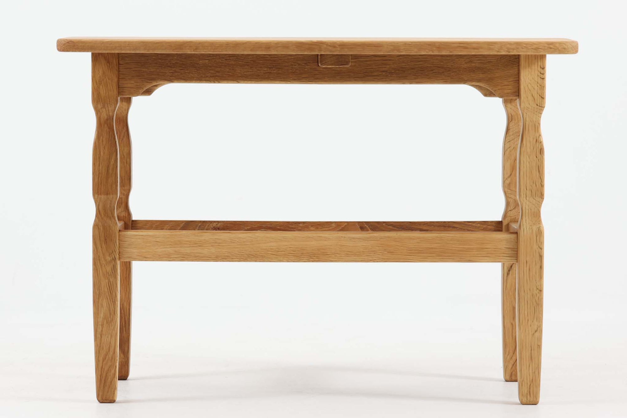 デンマークの建築家Henning Kjaernulf（ヘニング・ケアヌルフ）によってデザインされた、オーク無垢材のドロップリーフ式センターテーブルです。製造は高品質な家具で知られるEGモブラー社。無垢材ならではの美しい木目と、クラシカルで彫刻的な脚部のラインが印象的です。天板は両側に開閉式のリーフが備わっており、来客時や作業時には広げてスペースを拡張できるなど、実用性も兼ね備えています。高さ約51cmとやや高めの設計で、ソファ前に置いても手元が使いやすく、センターテーブルとして快適にお使いいただけます。必要に応じてサイドテーブルやティーテーブルとしても活躍する、用途の広い一台です。