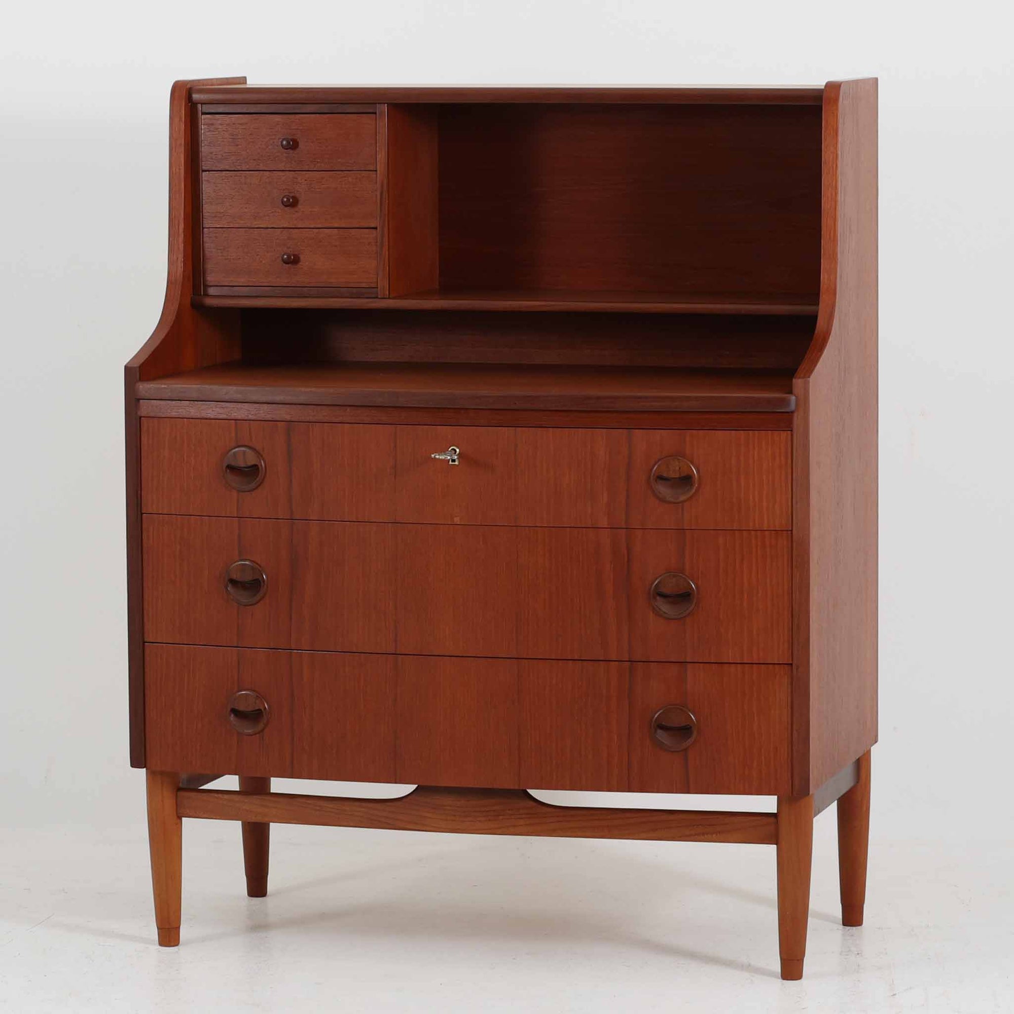 デンマーク製ライティングビューロです。上段には3杯の小引き出しとオープンスペースを備え、文具や手紙などを整理しながら使える実用的な設計です。丸い意匠の取っ手や滑らかな脚部のラインが北欧らしい温かみを感じさせ、チーク材の艶やかな木目が上品な印象を与えます。天板を開くと小さな書斎のような空間が広がり、リビングや寝室でも場所を取らず快適に作業が可能です。下段には3杯の引き出しを配置し、書類やノート類もしっかり収納できます。細身の脚部が軽やかさを添え、省スペースながら十分な収納力を兼ね備えた機能美あふれる一台です。経年によって増した風合いが魅力の、上質な北欧ビンテージ家具です。