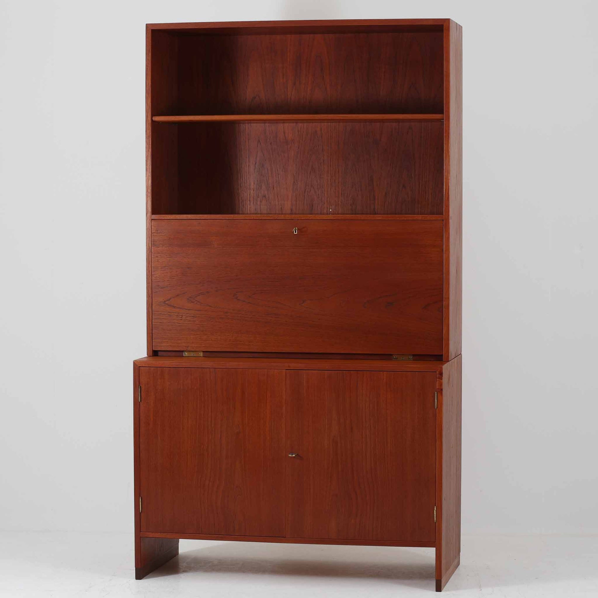 Hans J. Wegnerがデザインし、Ry Mobler社より製造されたこちらのディスプレイシェルフは、チーク材の上質な素材使いと真鍮パーツのディテールが際立つ北欧ビンテージ家具です。シンプルな直線的フォルムの中に、扉付き収納、ドロップダウン式のビューロスペース、小引き出しや仕切り棚など機能的な構造が備わっており、書斎でのデスク兼収納やリビングの飾り棚として多用途に活躍します。年月を経て深みを増した木肌が空間に落ち着いた存在感を与える逸品です。