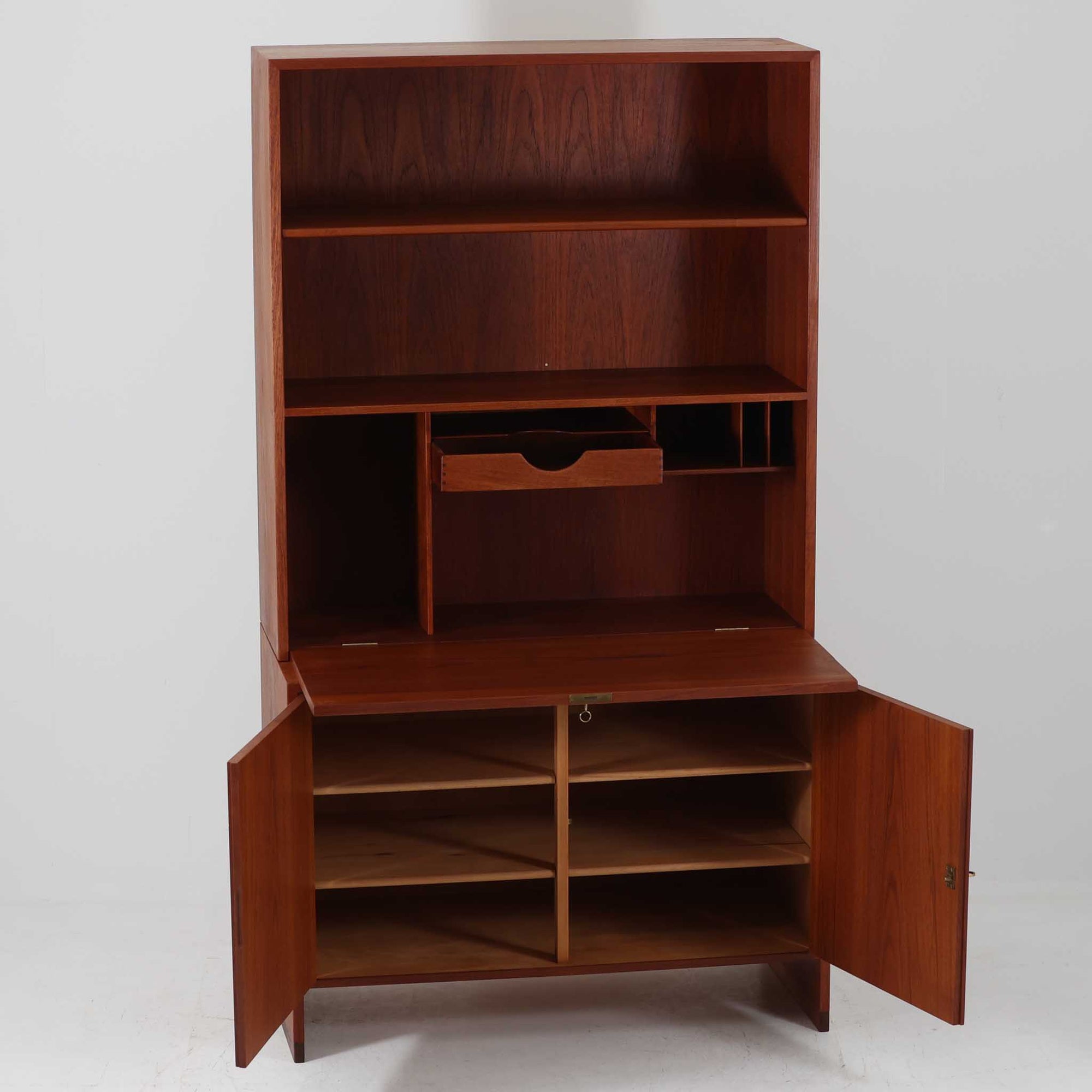 Hans J. Wegnerがデザインし、Ry Mobler社より製造されたこちらのディスプレイシェルフは、チーク材の上質な素材使いと真鍮パーツのディテールが際立つ北欧ビンテージ家具です。シンプルな直線的フォルムの中に、扉付き収納、ドロップダウン式のビューロスペース、小引き出しや仕切り棚など機能的な構造が備わっており、書斎でのデスク兼収納やリビングの飾り棚として多用途に活躍します。年月を経て深みを増した木肌が空間に落ち着いた存在感を与える逸品です。