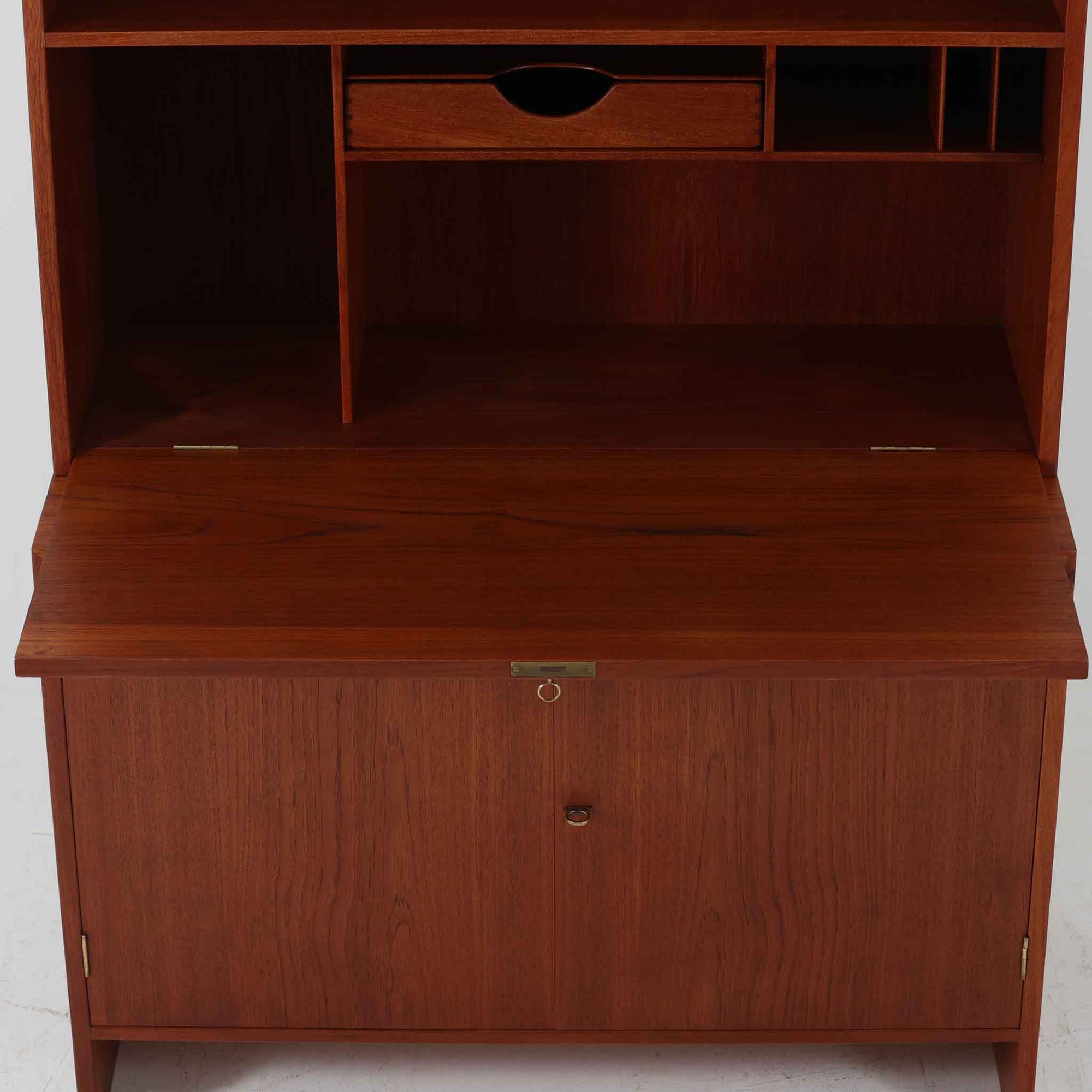 Hans J. Wegnerがデザインし、Ry Mobler社より製造されたこちらのディスプレイシェルフは、チーク材の上質な素材使いと真鍮パーツのディテールが際立つ北欧ビンテージ家具です。シンプルな直線的フォルムの中に、扉付き収納、ドロップダウン式のビューロスペース、小引き出しや仕切り棚など機能的な構造が備わっており、書斎でのデスク兼収納やリビングの飾り棚として多用途に活躍します。年月を経て深みを増した木肌が空間に落ち着いた存在感を与える逸品です。