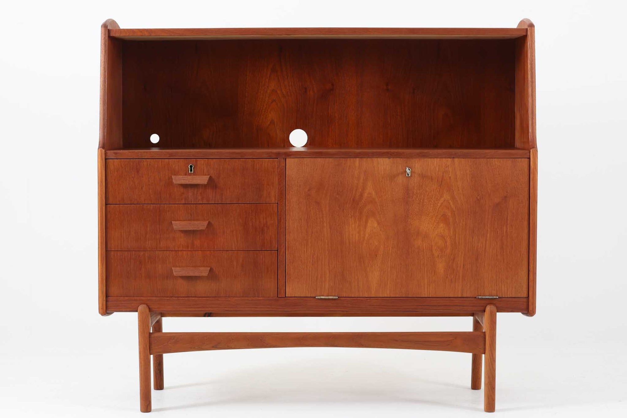 北欧デンマークで製造されたチーク材のビンテージライティングビューローです。シンプルな直線と柔らかな曲面が調和した洗練された佇まいで、北欧ミッドセンチュリーらしい温もりと機能美が感じられます。上部はオープンシェルフになっており、背板には前オーナーによって開けられた配線用のコードホールも備えているため、ルーターやオーディオ機器の収納にも便利です。下部には3杯の引き出しと、鍵付きの開き扉収納が備わっており、扉を開けると棚板が設けられた実用的な構造です。特にビューローにこのような「開き扉」を採用したデザインは珍しく、収納力と個性を兼ね備えた一台となっています。奥行は約37cmと圧迫感が少なく、リビングやワークスペースなど幅広い空間に取り入れやすいサイズ感です。