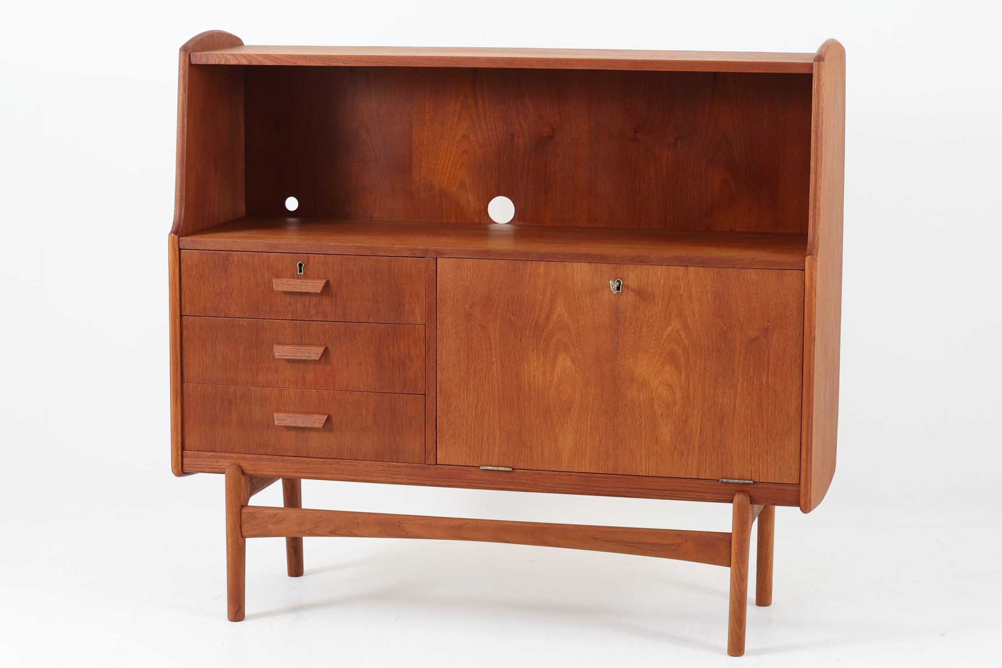 北欧デンマークで製造されたチーク材のビンテージライティングビューローです。シンプルな直線と柔らかな曲面が調和した洗練された佇まいで、北欧ミッドセンチュリーらしい温もりと機能美が感じられます。上部はオープンシェルフになっており、背板には前オーナーによって開けられた配線用のコードホールも備えているため、ルーターやオーディオ機器の収納にも便利です。下部には3杯の引き出しと、鍵付きの開き扉収納が備わっており、扉を開けると棚板が設けられた実用的な構造です。特にビューローにこのような「開き扉」を採用したデザインは珍しく、収納力と個性を兼ね備えた一台となっています。奥行は約37cmと圧迫感が少なく、リビングやワークスペースなど幅広い空間に取り入れやすいサイズ感です。