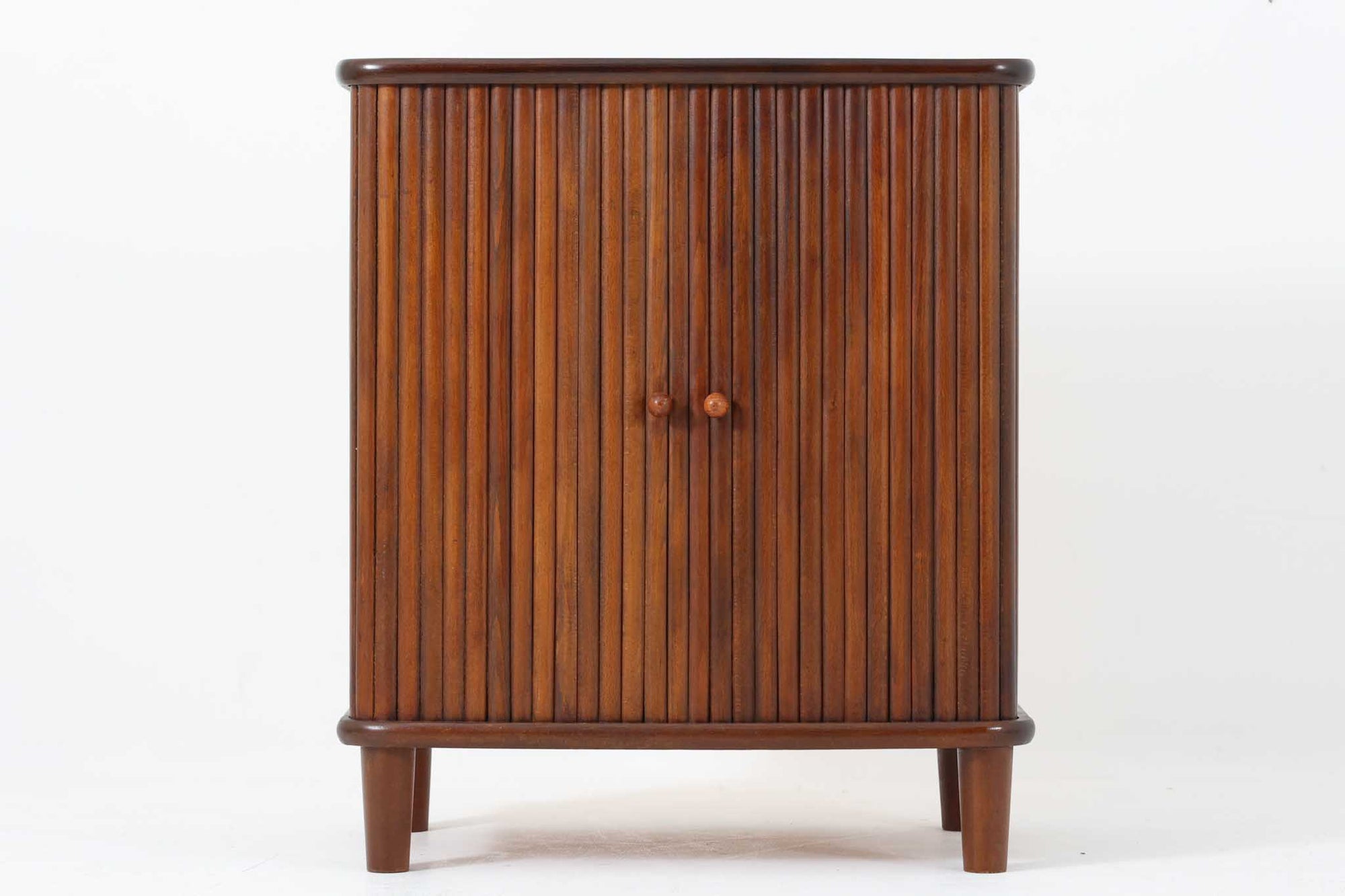 北欧デンマークで製造された、蛇腹扉が特徴的なビンテージキャビネットです。素材にはビーチ材と思われる無垢材や突板が使用されており、ステイン塗装により落ち着いたブラウンカラーに仕上げられています。前面には細かな縦ラインが連続する蛇腹扉が配され、開閉時に滑らかに動く美しい構造が魅力です。内部には棚板が一枚付属しており、収納としての機能性も十分です。丸脚や小さな丸い取っ手も北欧らしい柔らかなディテールで、リビングや玄関、寝室などさまざまな空間に自然に溶け込む一台です。