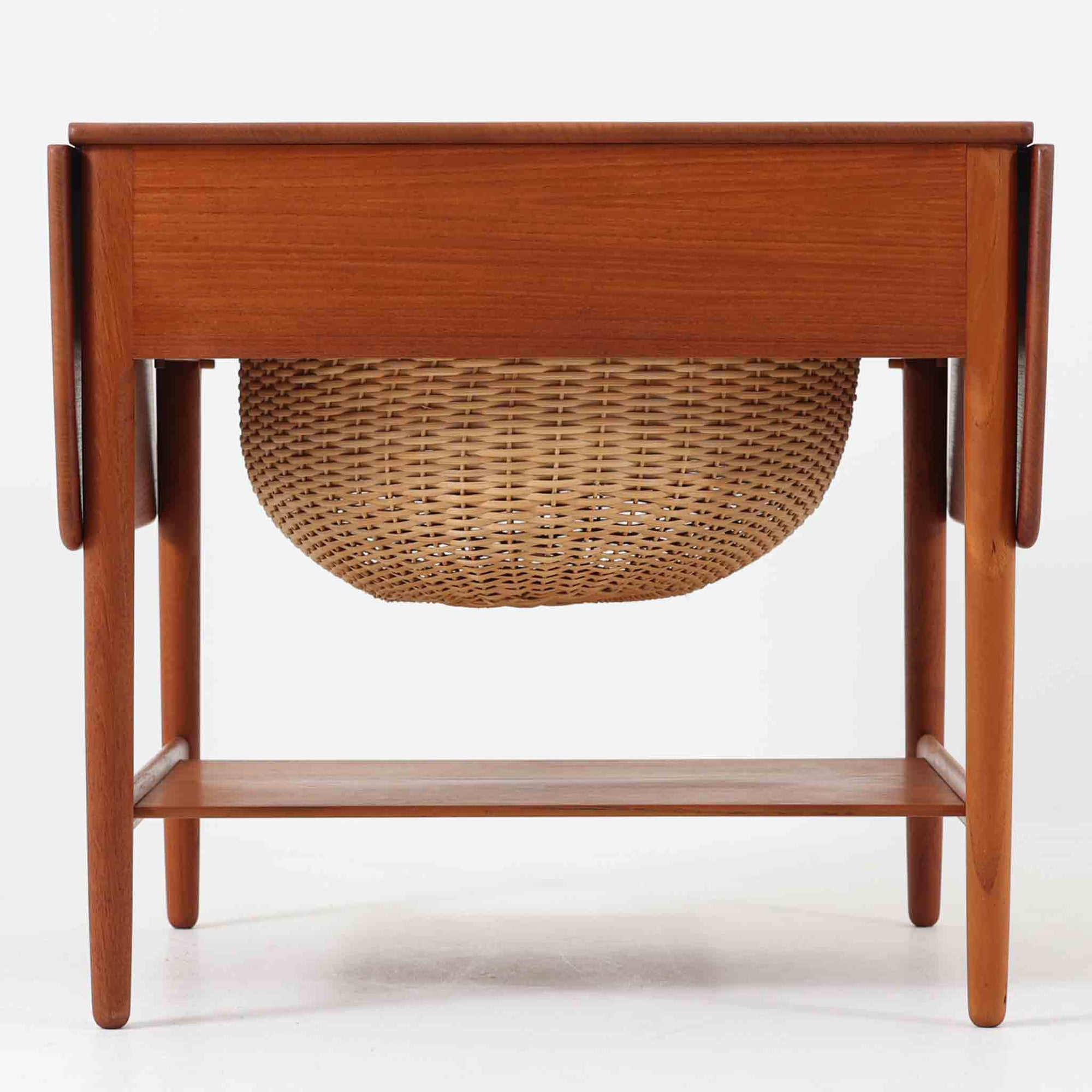 Hans J. Wegnerがデザインし、Andreas Tuck社から発表されたAT33ソーイングテーブルは、チーク無垢材を贅沢に用いた希少なモデルです。両側のバタフライ式天板で作業スペースを拡張でき、仕切り付きの引き出しとラタン製バスケットが裁縫道具や小物を美しく収納します。下段の棚板も備えた実用性の高い構造で、繊細な造作と職人技が光る北欧ビンテージ家具の逸品です。