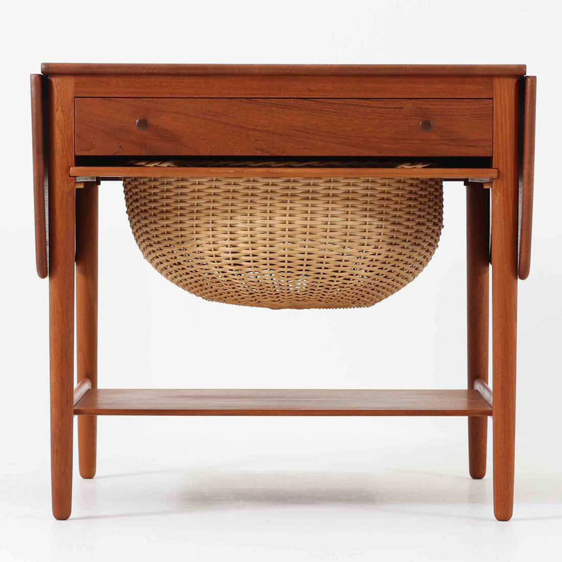 Hans J. Wegnerがデザインし、Andreas Tuck社から発表されたAT33ソーイングテーブルは、チーク無垢材を贅沢に用いた希少なモデルです。両側のバタフライ式天板で作業スペースを拡張でき、仕切り付きの引き出しとラタン製バスケットが裁縫道具や小物を美しく収納します。下段の棚板も備えた実用性の高い構造で、繊細な造作と職人技が光る北欧ビンテージ家具の逸品です。