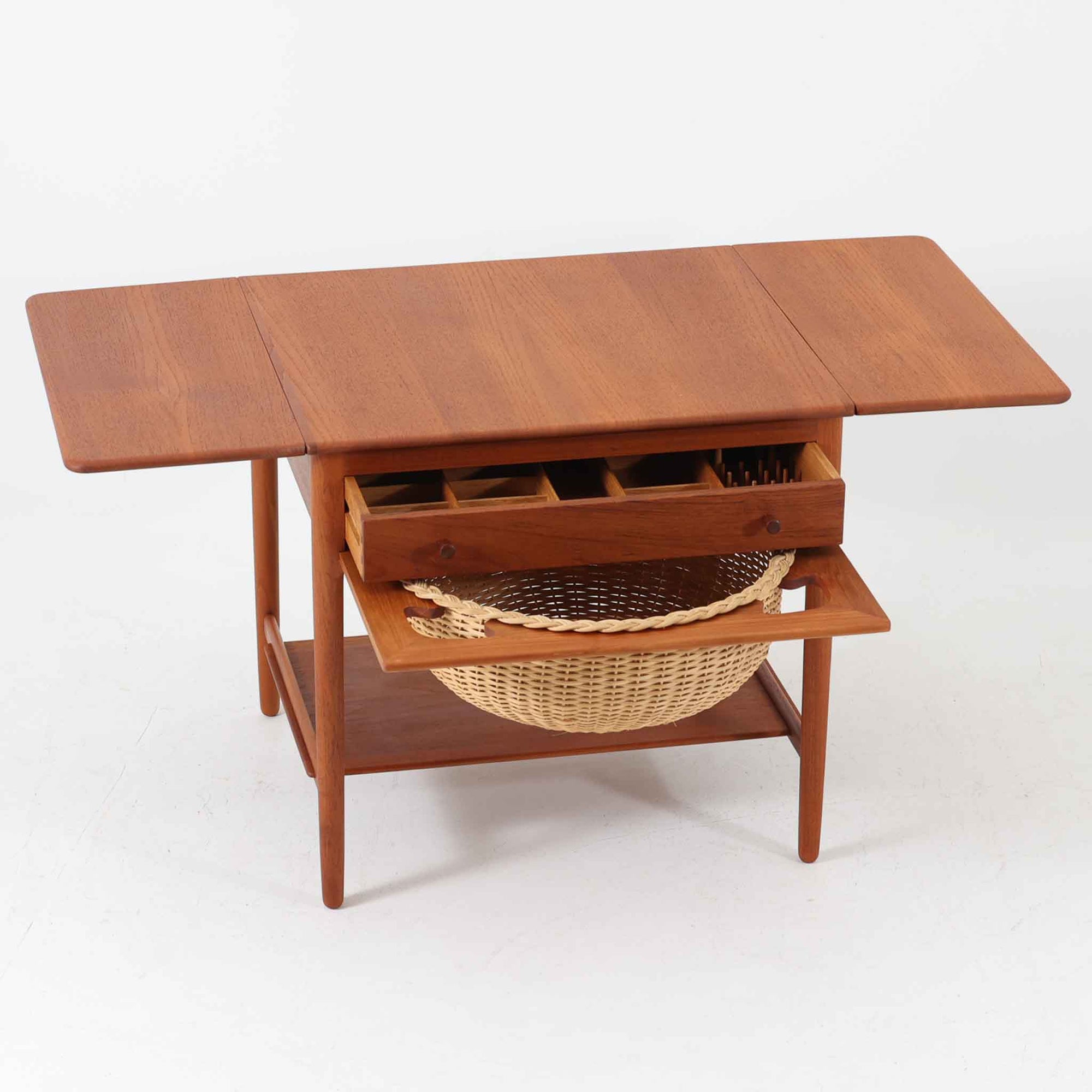 Hans J. Wegnerがデザインし、Andreas Tuck社から発表されたAT33ソーイングテーブルは、チーク無垢材を贅沢に用いた希少なモデルです。両側のバタフライ式天板で作業スペースを拡張でき、仕切り付きの引き出しとラタン製バスケットが裁縫道具や小物を美しく収納します。下段の棚板も備えた実用性の高い構造で、繊細な造作と職人技が光る北欧ビンテージ家具の逸品です。