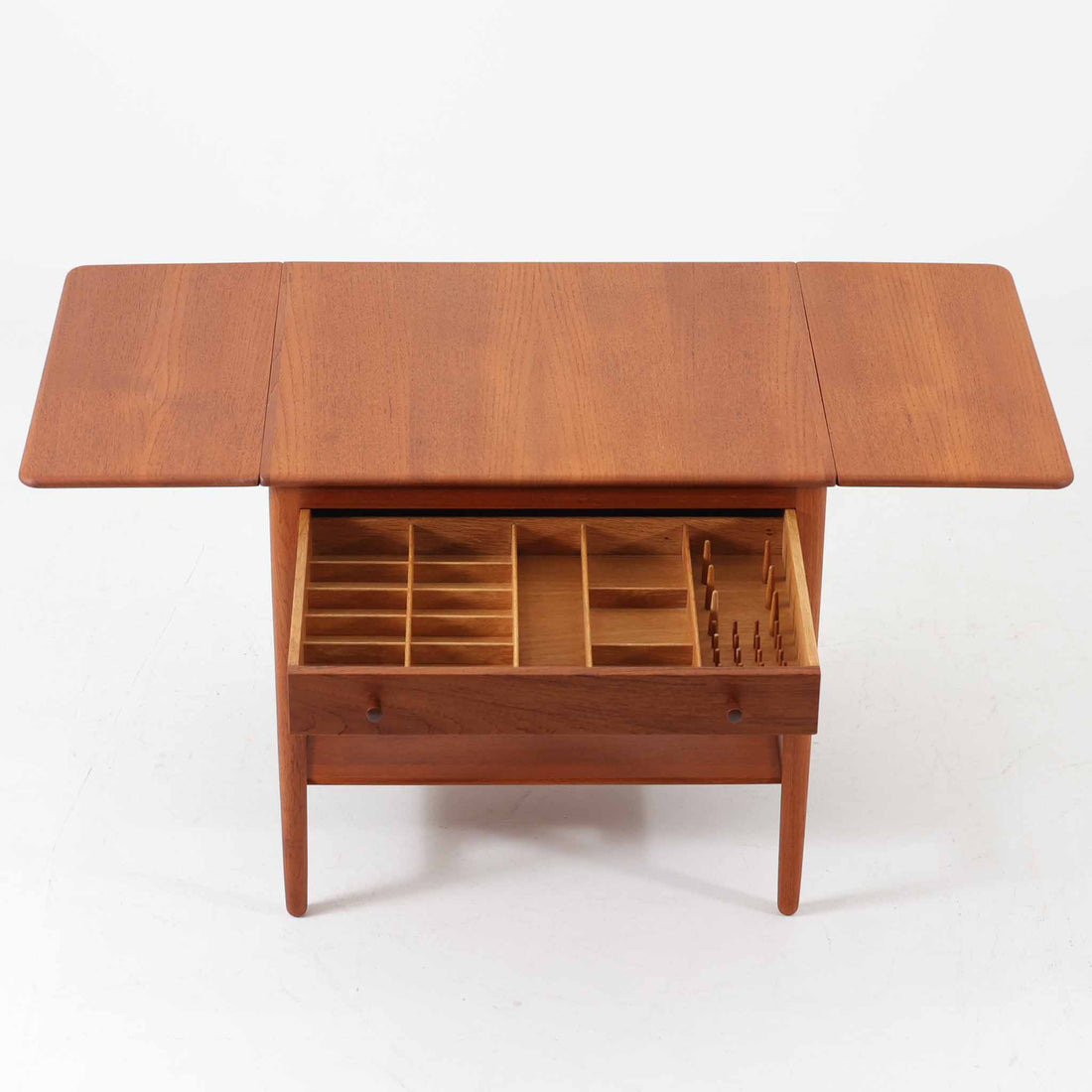 Hans J. Wegnerがデザインし、Andreas Tuck社から発表されたAT33ソーイングテーブルは、チーク無垢材を贅沢に用いた希少なモデルです。両側のバタフライ式天板で作業スペースを拡張でき、仕切り付きの引き出しとラタン製バスケットが裁縫道具や小物を美しく収納します。下段の棚板も備えた実用性の高い構造で、繊細な造作と職人技が光る北欧ビンテージ家具の逸品です。