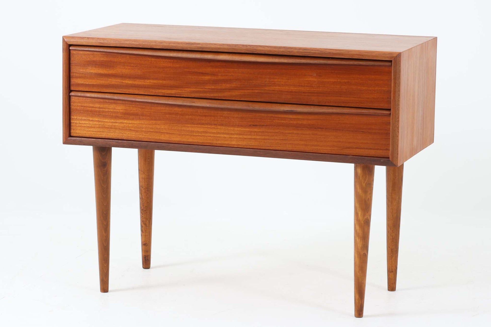 温かみのある木目が魅力的なチーク材を使用した、北欧ビンテージの2段チェストです。直線的なフォルムに、先細りのナチュラルな色味のビーチ材テーパー脚を組み合わせたミニマルなデザインが美しく、空間に軽やかな印象をもたらします。引き出し前板は、手掛けによって開閉できる仕様となっており、すっきりとしたデザイン性と実用性を兼ね備えています。フレーム縁も丁寧に削り出されており、細部までクオリティの高い仕上がりが感じられます。底面には「Jacob Amstrup Funder」の刻印が確認でき、かつてデンマーク・VestbjergにあるRenovere møblerにて丁寧に修復されたことがうかがえます。ベッドサイドやリビングの一角、玄関収納など、さまざまなシーンで活躍する一台です。